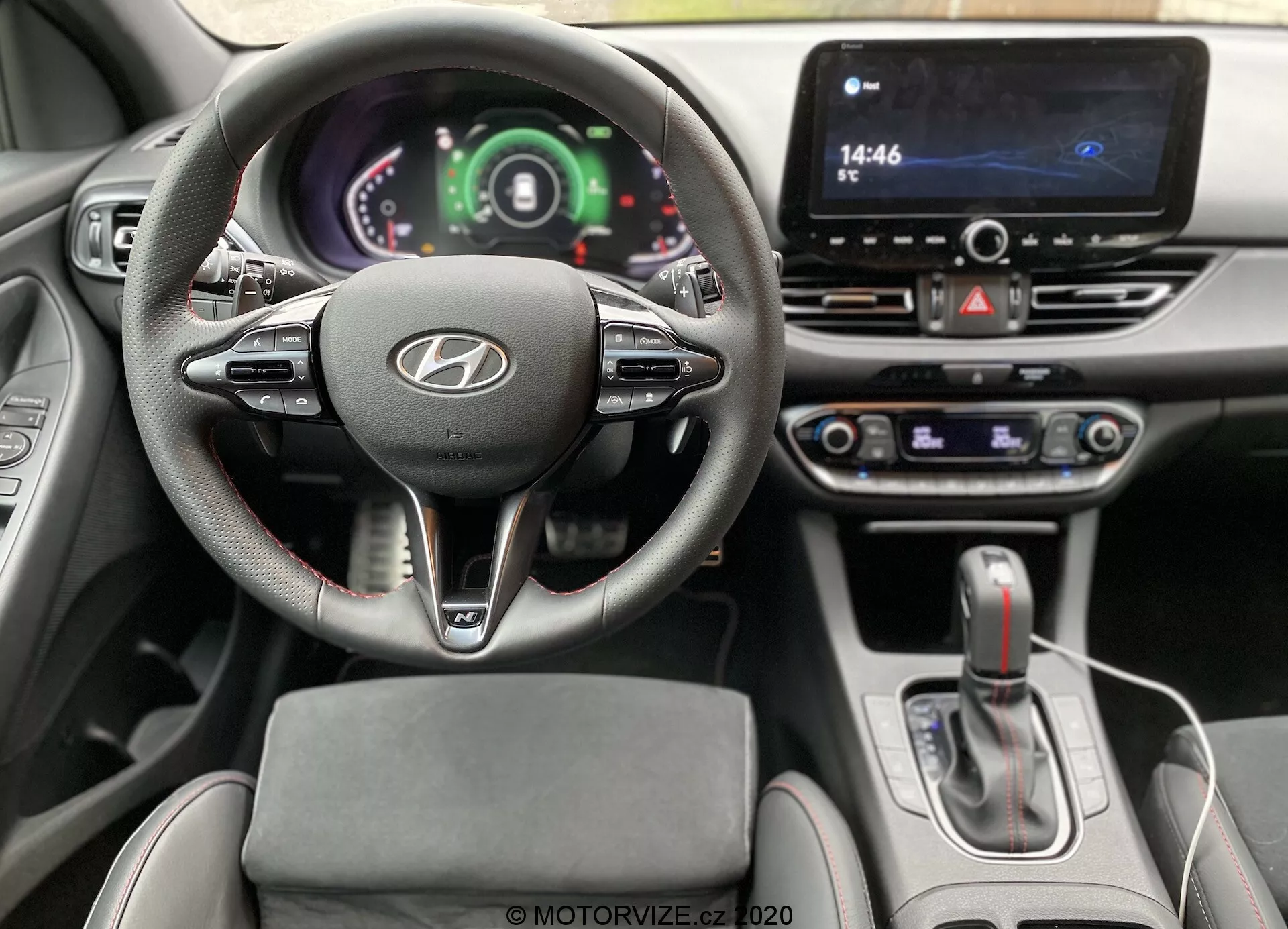 Pohľad na interiér z pohľadu vodiča vnútri Hyundai i30 2020 (i30-kombi), ktorý ukazuje volant s logom Hyundai, montovanými ovládačmi a digitálnym prístrojovým štítom. Viditeľné sú dotykový infotainment systém, volič automatickej prevodovky a ovládacie prvky klimatizácie, čo naznačuje pokročilú technológiu a moderný dizajn.