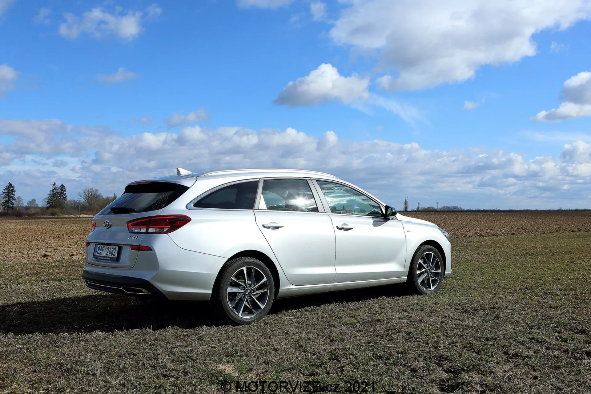 Detailný pohľad na biely Hyundai i30 kombi 2020 zaparkovaný na trávnatej lúke s čiastočne zatiahnutou oblohou v pozadí. Obrázok je zhotovený z trojštvrtinového zadného uhla, ukazujúci dizajnové prvky ako moderné zadné svetlá obkresľujúce boky vozidla, zadné okno s stieračom, malý strešný spojler, viacramenné zliatinové kolesá a bočné zrkadlo s možnými modernými funkciami. Viditeľné sú medzery zadných dvier pre cestujúcich a dvier batožinového priestoru, čo naznačuje minimálne štyri dvere. Hoci výfuk nie je výrazne viditeľný, pôsobí nenápadne. Auto je ostro zaostrené, zvýrazňujúc dizajn a funkcie, so základným prostredím poskytujúcim kontextovú kulisu.
