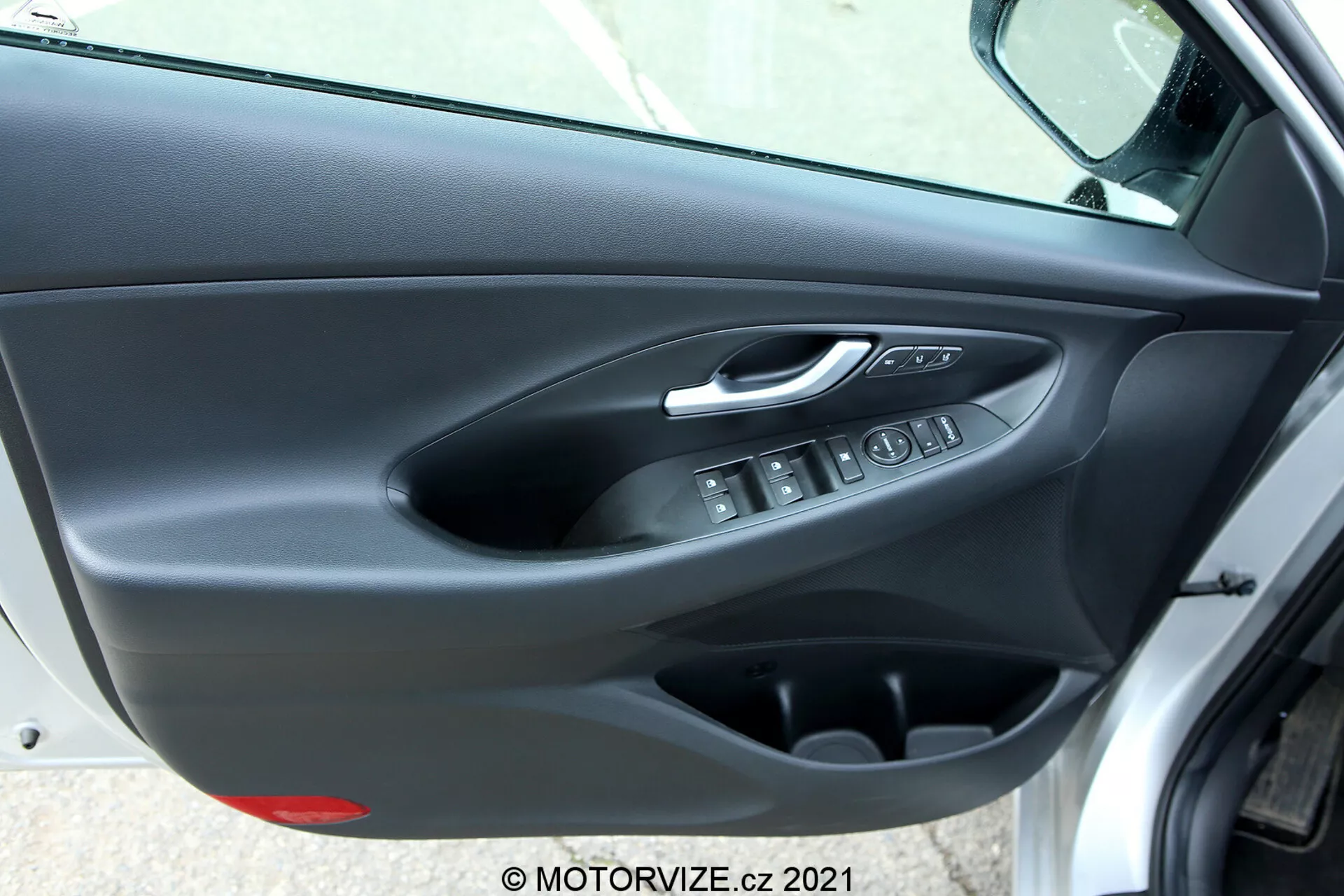 Vnútorný panel dverí Hyundai i30 2020 (i30-kombi) z pohľadu šikmého uhla, ukazujúci striebornú chrómovanú kľučku dverí, ergonomické tlačidlá na ovládanie elektrických okien, elektronické tlačidlo zámku dverí, úložné vrecko a dvojtextúrný dverný obklad s mäkkým horným panelom a tuhým plastovým dolným dielom v tmavých farbách.