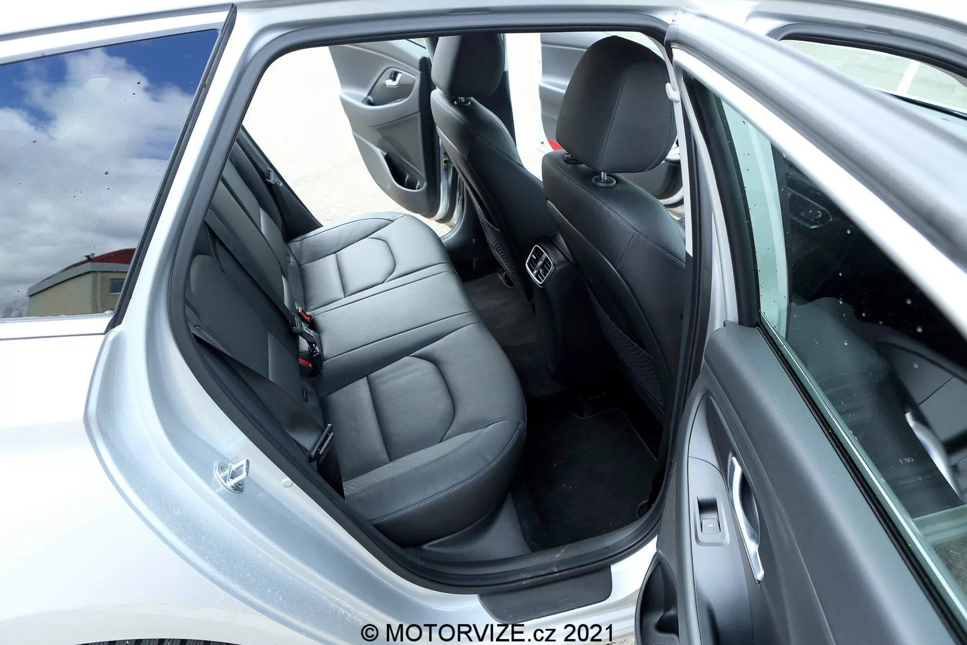Detailný pohľad na zadnú časť interiéru Hyundai i30 2020. Obrázok zobrazuje otvorené zadné dvere s jasným pohľadom na zadné sedadlá z tmavej kože, ktoré majú individuálne opierky hlavy a bezpečnostné pásy. Panel dverí obsahuje sivé alebo strieborné akcenty s ovládacími tlačidlami a mriežkou reproduktora, indikujúci technologické a audio funkcie. Viditeľné sú predné sedadlá a B-stĺpik, odrážajúc podobný dizajn a materiál ako zadné sedadlá. Interiér, prevažne čierny s niektorými striebornými akcentmi, je dobre udržiavaný, so zameraním na pohodlie a dizajn.