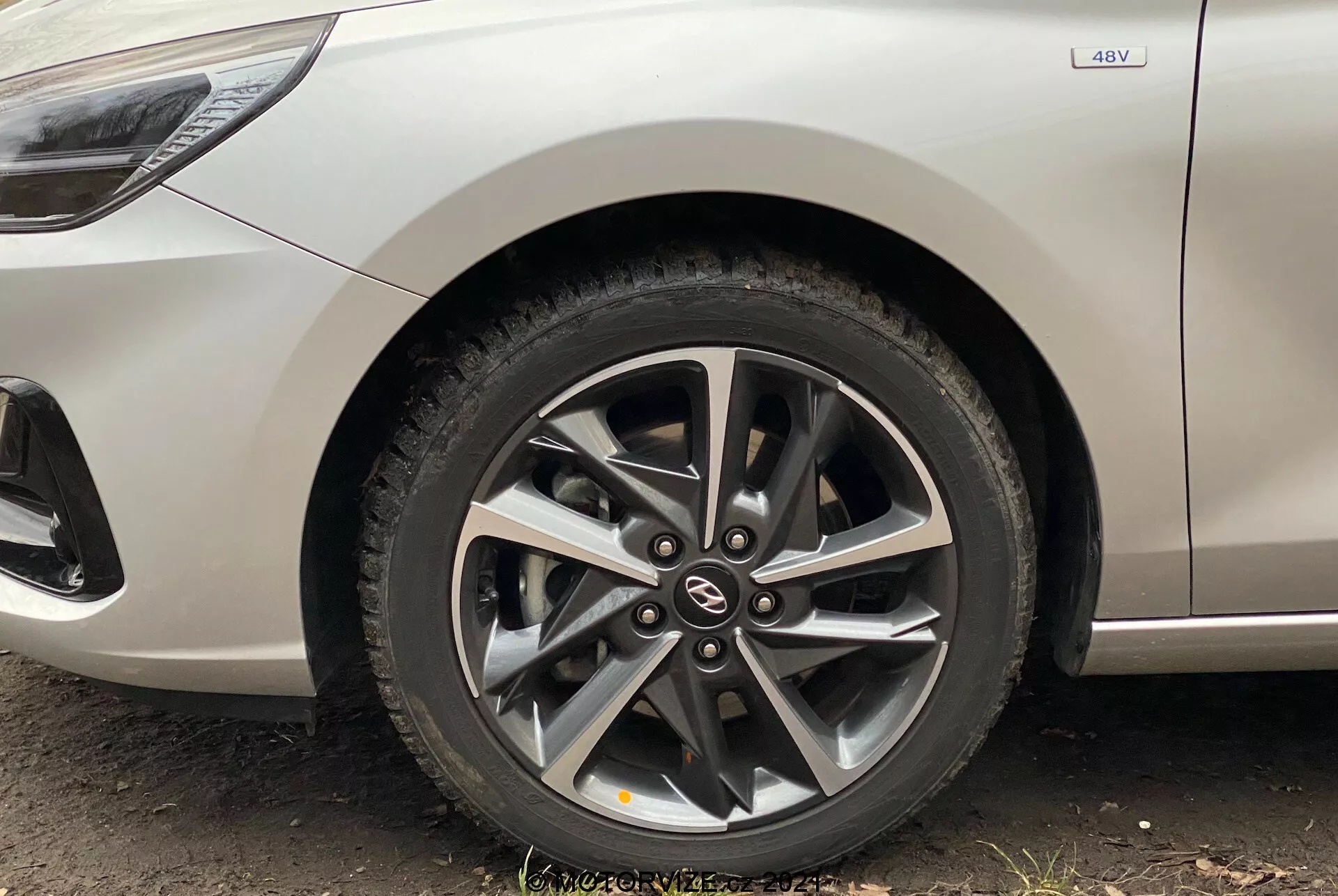 Záber na predné koleso Hyundai i30 Kombi 2020, zobrazujúci moderné hliníkové koleso s viacramenným dizajnom a kotúčovými brzdami viditeľnými cez rámy. Pohľad, zaznamenaný z nízkeho uhla takmer pri zemi, zahŕňa časť bočnej karosérie v metalickej striebornej farbe, s viditeľným odznakom 4WD naznačujúcim schopnosť pohonu všetkých štyroch kolies. Obraz zdôrazňuje čistý dizajn auta so zakrivenými líniami a drobnými charakterovými detailmi nad oblúkom kolesa.