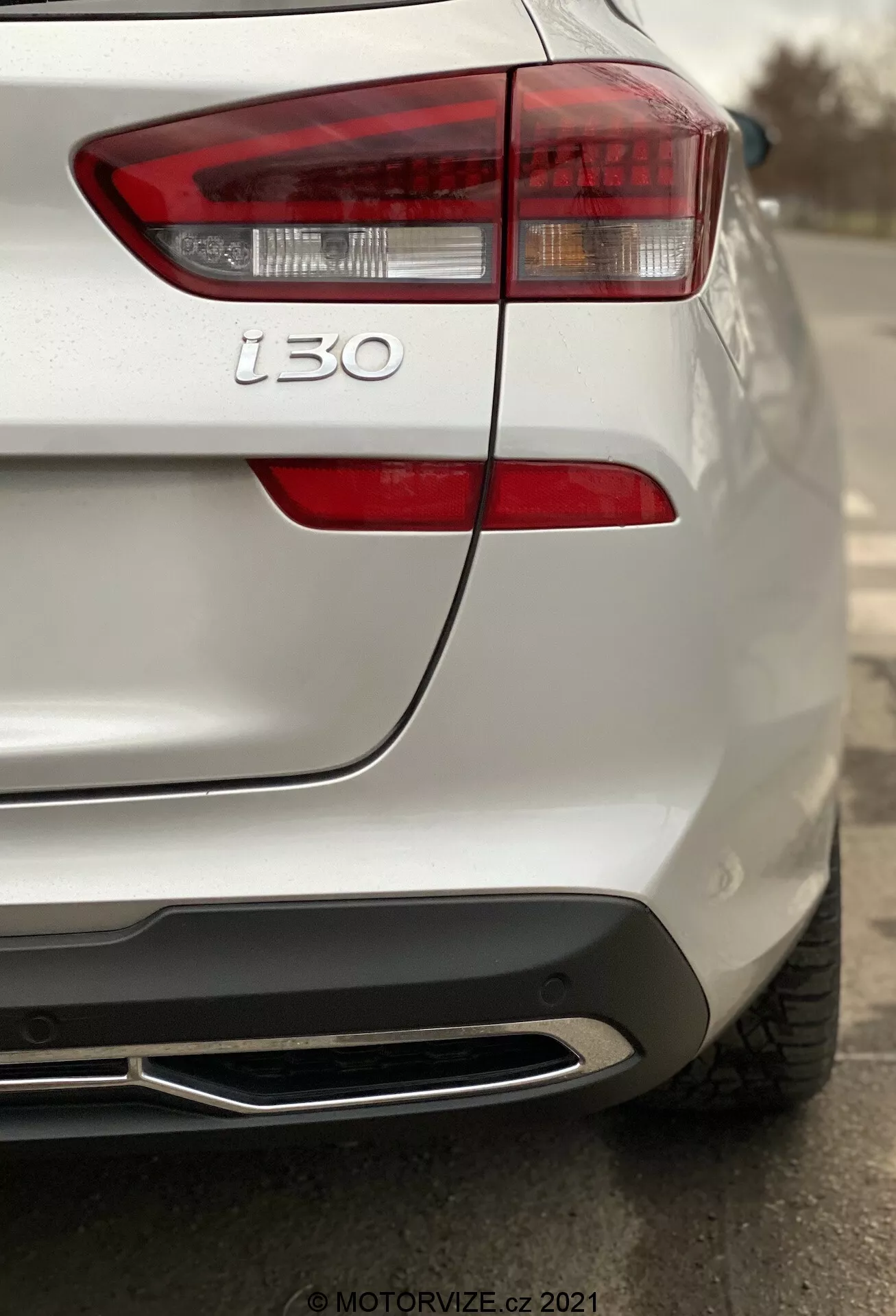 Detailný zadný pohľad na biely Hyundai i30 2020 (Kombi) so zameraním na ľavé zadné svetlo a ľavý zadný štvrťový diel, prezentujúci moderné dizajnové línie, čisté brzdové svetlo, smerovky, odznak i30, detaily textúry laku, parkovací senzor, integrovaný dizajn a chrómovanú koncovku výfuku s malou hĺbkou ostrosti zvýrazňujúcou zadný dizajn auta.