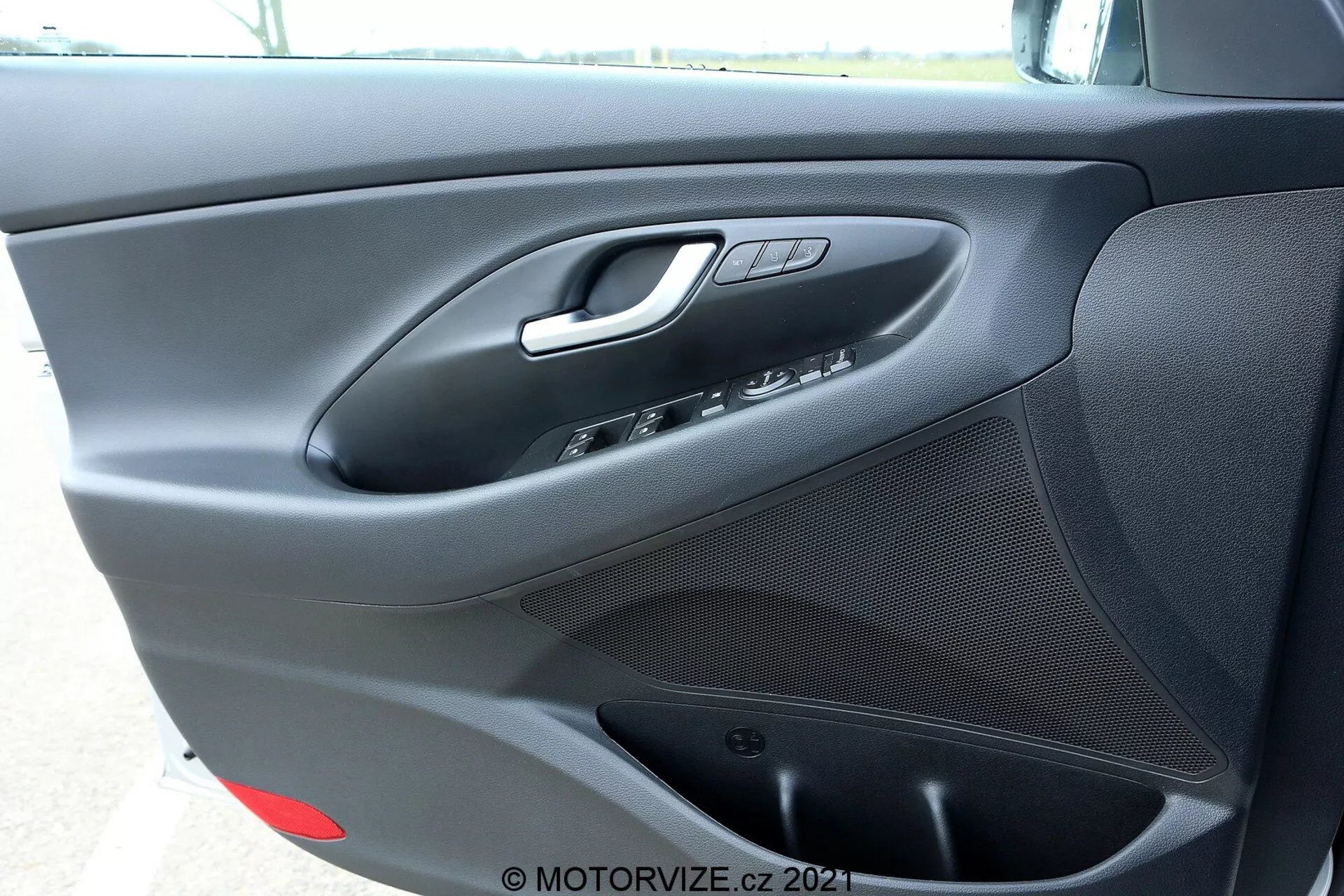 Záber na vnútorný dverový panel na strane vodiča Hyundai i30 Kombi 2020, ukazujúci striebornú kľučku dverí, ovládanie okien a zrkadiel, tlačidlo zámku dverí, integrovaný reproduktor v spodnej časti a textúrovaný matný čierny povrch s pravdepodobne malým úložným priestorom na spodnej časti.