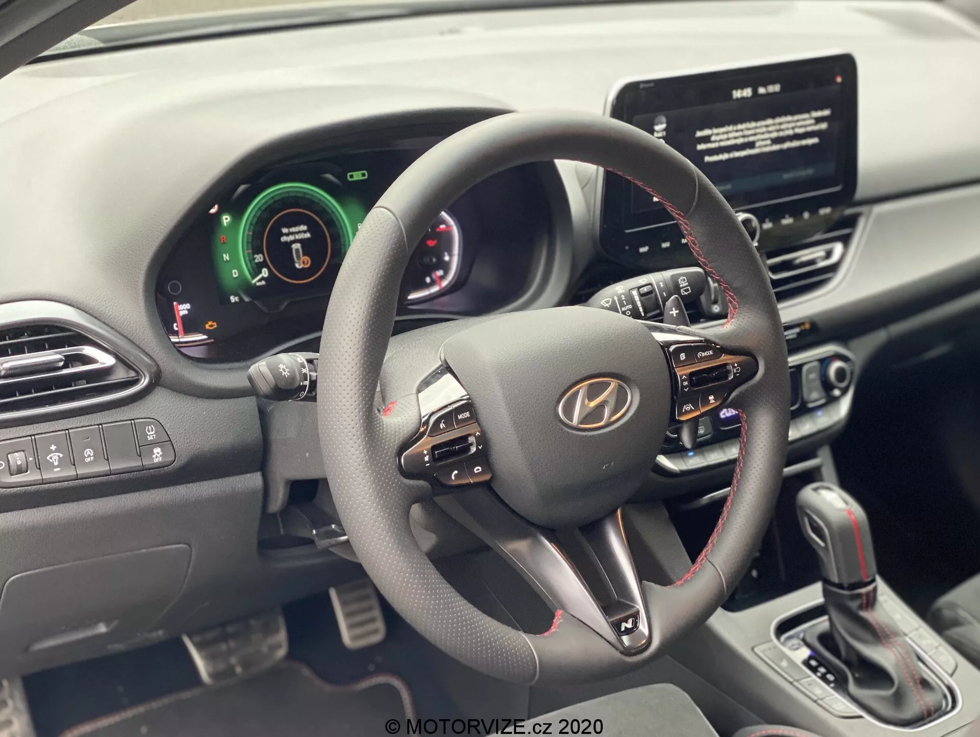 Pohľad do interiéru Hyundai i30 2020 (i30-kombi) z miesta vodiča, zameraný na volant a prístrojovú dosku. Moderný volant má niekoľko ovládacích tlačidiel pre hlasitosť, hlasové príkazy a hands-free volanie. Palubná doska kombinuje digitálne a analógové prvky, čo signalizuje pokročilé asistenčné systémy vodiča. Viditeľná je centrálná dotyková obrazovka infotainmentu, ktorá naznačuje navigáciu a prehrávanie médií. Stredová konzola ukazuje tlačidlá klimatizácie a radiacu páku automatickej prevodovky. Ovládanie okien a spätných zrkadiel je na vodičových dverách.