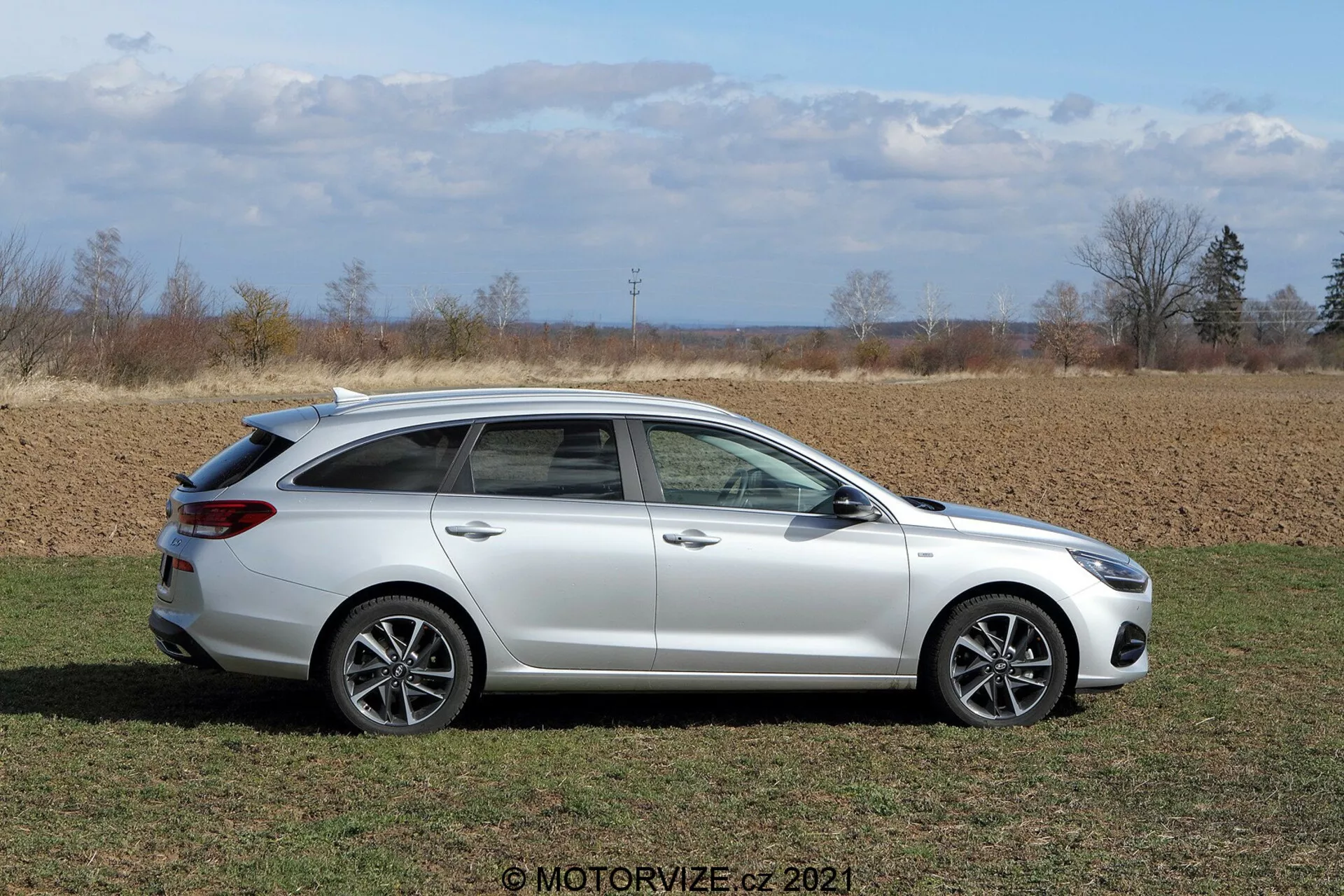 Bočný profil bieleho Hyundai i30 2020 kombi (i30-kombi) zaparkovaného na vidieku s poľnohospodárskymi pozemkami v pozadí. Viditeľný je elegantný dizajn svetlometov, viacramenné zliatinové kolesá, bočné zrkadlá s integrovanými smerovými svetlami a strešný spojler v jasnom, prirodzenom dennom svetle.