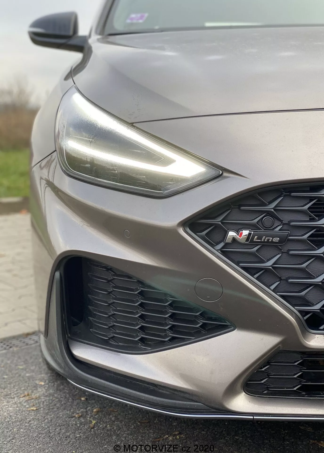 Zblízka, uhlový pohľad zameraný na prednú ľavú stranu sivého Hyundai i30 2020 N Line. Obrázok zdôrazňuje športový dizajn vrátane jemného obrysu kapoty, ostrých LED svetlometov, mriežky v tvare včelieho plástu s výrazným červeným odznakom 'N Line' a zložitého nárazníka s pokročilými senzormi naznačujúcimi moderné asistenčné systémy pre vodiča.