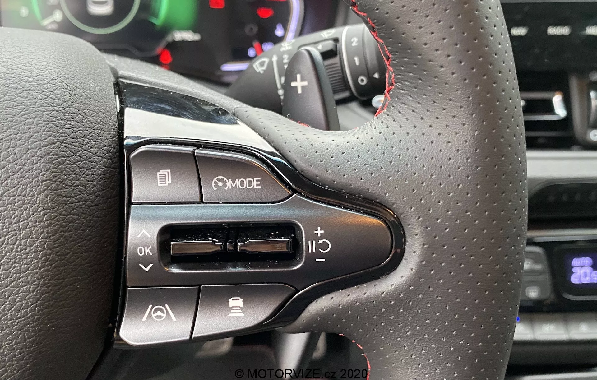 Detailný pohľad na ľavú stranu volantu Hyundai i30 Kombi 2020 s textúrou podobnou koži a červeným prešívaním, moderné multifunkčné ovládače, vrátane tlačidla MODE, navigačnej klávesnice, ovládania hlasitosti a čiastočný pohľad na radiacu páku. V pozadí je nejasná prístrojová doska alebo ukazovatele z pohľadu vodiča.