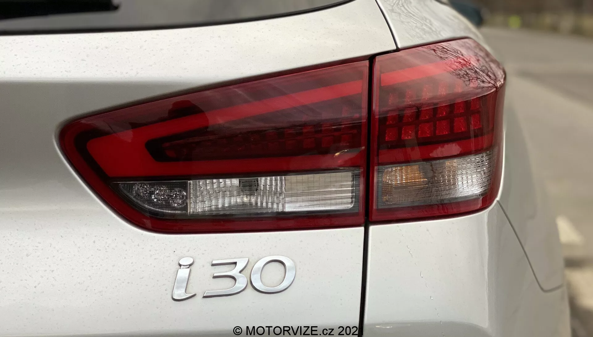 Detailný pohľad na zadnú ľavú stranu Hyundai i30 Kombi 2020 so zameraním na moderné zadné svetlo so zreteľnými oddeleniami medzi červeným brzdovým svetlom, bielym spätným svetlom a oranžovým indikátorom podľa európskych špecifikácií. Záber zahŕňa časť veka batožinového priestoru, zadného nárazníka a emblém 'i30', pravdepodobne so strieborným alebo bielym lakom. Malá hĺbka ostrosti zdôrazňuje zadné svetlo a emblém, s rozmazaným pozadím a viditeľným vodoznakom 'MOTORVIZE.cz 202'.