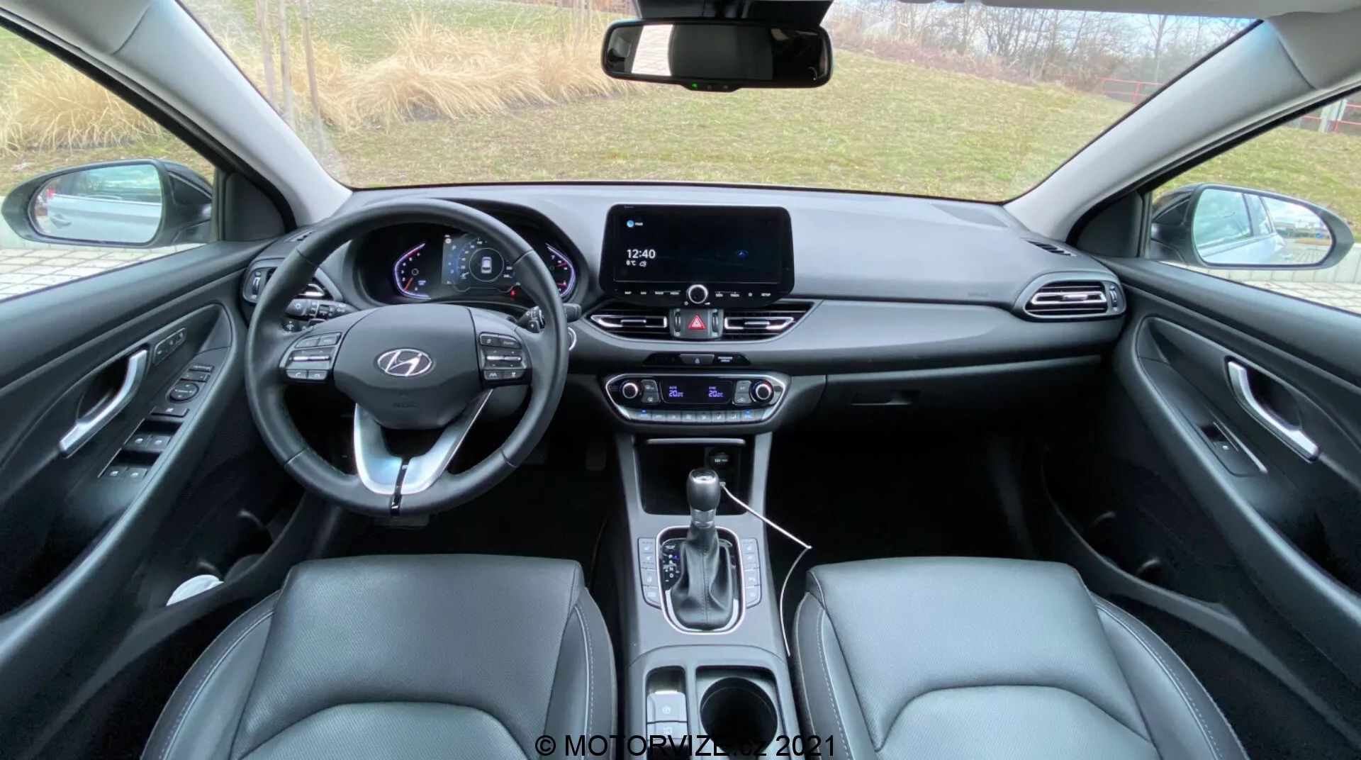 Pohľad do interiéru Hyundai i30 2020 (i30-Kombi) zo zadného sedadla, ktorý ukazuje multifunkčný volant s ovládacími prvkami, digitálny prístrojový panel indikuje parkovací režim a otvorené dvere vodiča, dotykový infotainment systém, automatickú radiacu páku, predné sedadlá čalúnené kožou, podrobné dverné panely s elektrickými ovládateľnými prvkami, rozmanité vnútorné materiály vrátane syntetickej kože a kovových prvkov, hornú konzolu s priestorom pre slnečné okuliare a tlačidlá, automaticky stmavené spätné zrkadlo s dodatočnými tlačidlami a čiastočne viditeľnými koberčekmi.