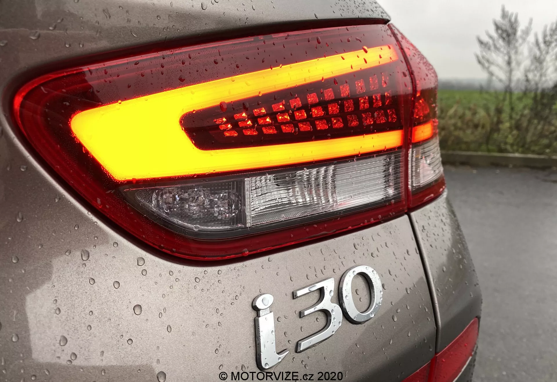 Detailný záber na zadnú časť vozidla Hyundai i30 2020 (i30-kombi) so zameraním na moderný LED zadný svetlomet vyžarujúci jasné oranžovo-žlté svetlo, ktoré naznačuje funkciu smerovky alebo núdzových svetiel. Fotografia zdôrazňuje nápis modelu i30 a kvapky vody na povrchu vozidla, čím ukazuje pokročilú technológiu automobilových svetiel a detailné prevedenie vozidla.