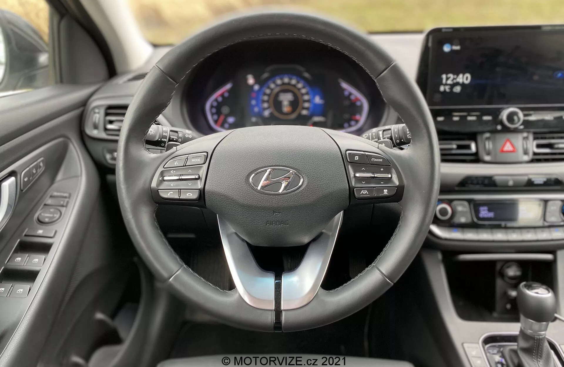 Vnútorný pohľad na Hyundai i30 Kombi 2020 z perspektívy vodiča, zobrazujúci moderný kokpit s trojramenným volantom, digitálnym prístrojovým panelom a dotykovým systémom infotainmentu. Volant obsahuje ovládacie tlačidlá pre audio, komunikáciu a tempomat s dvojfarebným dizajnom. Osvetlený prístrojový panel zobrazuje tachometer, otáčkomer a centrálny informačný displej. V strednej časti palubnej dosky sú vzduchové otvory a ovládanie klimatizácie. Čiastočný pohľad na radiacu páku naznačuje manuálnu prevodovku. Auto sa zdá byť v pokoji, bez rozmazania viditeľného cez okná.
