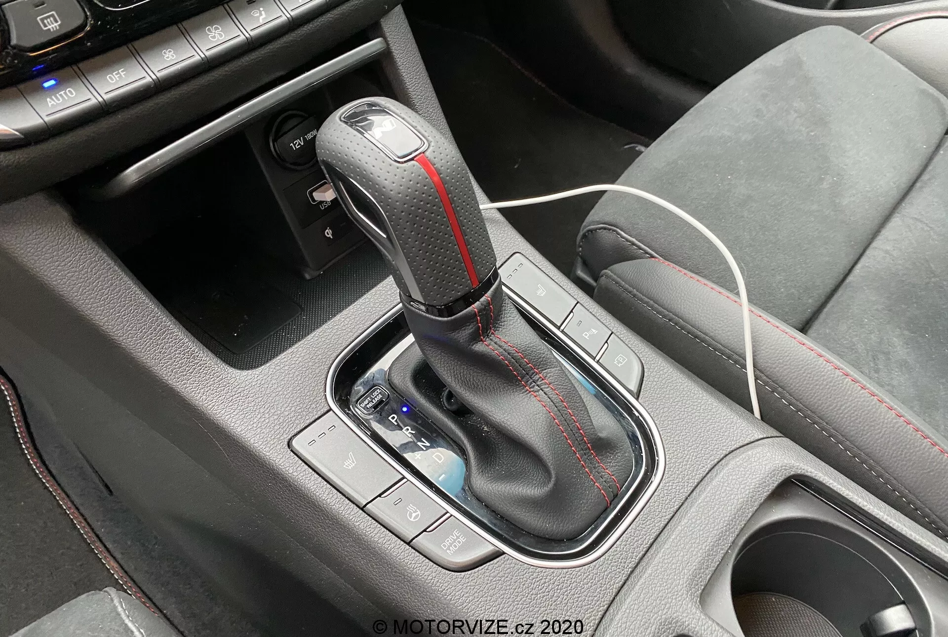 Detailný záber na stredovú konzolu Hyundai i30 2020 kombi, so zameraním na automatickú radiacu páku. Páka má koženú hlavicu s kovovými akcentmi a červeným kontrastným prešívaním. Pred radiacou pákou je viditeľný malý úložný priestor s bielym nabíjacím káblom, čo naznačuje prítomnosť USB portov alebo 12V zásuvky. Okolité konzolové plochy sú vyrobené z textúrovaného plastu pre lepšie uchopenie. Ovládacie prvky naľavo od páky, pravdepodobne pre jazdné režimy alebo kontrolu trakcie, sú mierne rozmazané.