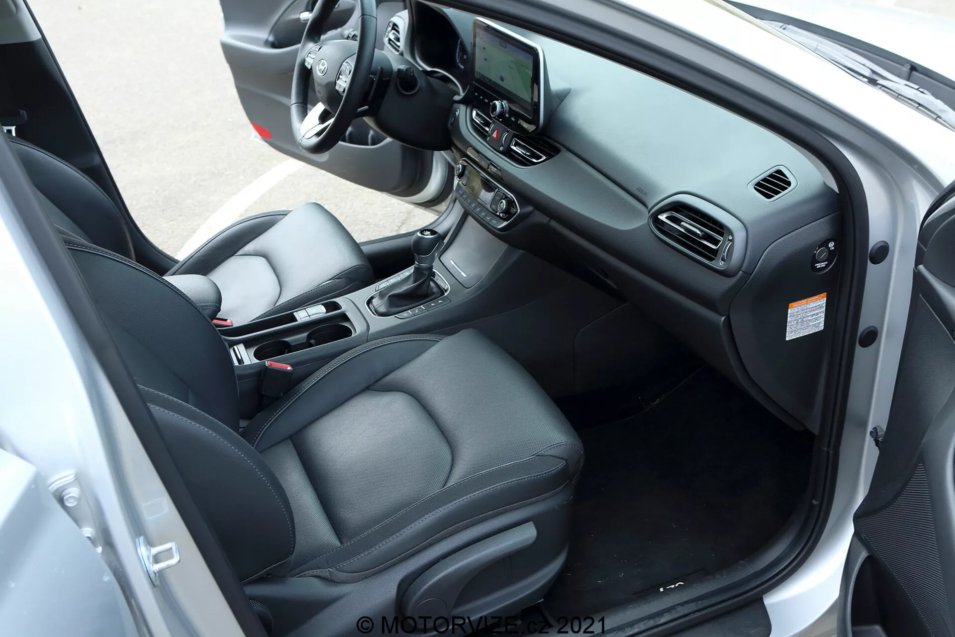 Interiér Hyundai i30 kombi 2020, zachytený z diagonálneho, mierne zvýšeného uhla zameriavajúceho sa na stranu vodiča. Obrázok zobrazuje kožou čalúnené sedadlo vodiča s elektronickými ovládacími prvkami, multifunkčný volant s rôznymi tlačidlami ovládania, centrálne dotykové infotainmentové zariadenie a automatickú radiacu páku na stredovej konzole. Ďalšie prvky zahŕňajú držiaky na poháre, úložný priestor, automatickú klimatizáciu a ovládanie elektrických okien a spätných zrkadiel na paneli dverí vodiča. Dvere auta sú široko otvorené, aby poskytli komplexný výhľad na prístrojovú dosku, usporiadanie sedadiel a technologické a komfortné prvky interiéru.