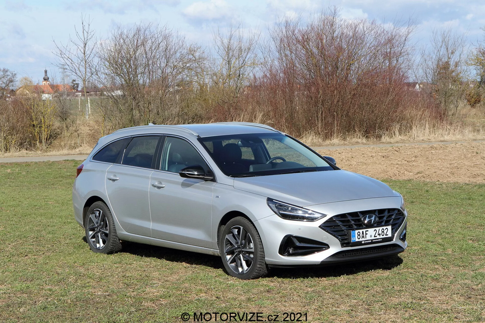 Trojštvrťový čelný pohľad na moderný hatchback Hyundai i30 2020 kombi, nasnímaný z vyvýšeného uhla, ktorý zvýrazňuje prednú mriežku s voštinovým dizajnom, elegantné svetlomety, viacpruhové zliatinové kolesá, bočné zrkadlá so zabudovanými smerovkami a strešné koľajnice na kovovom tele s hladkými, tvarovanými líniami v slnečnom prírodnom prostredí.