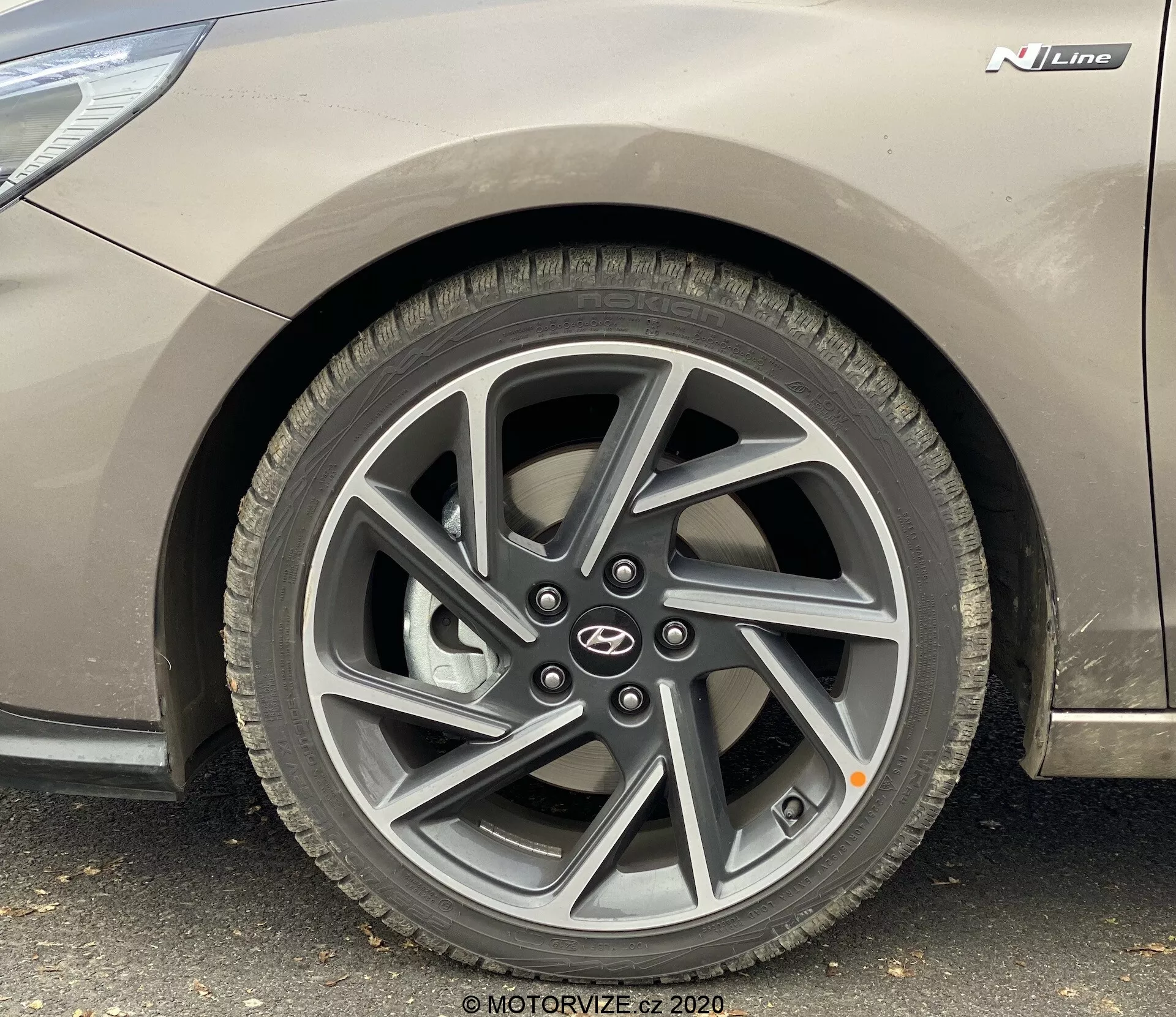 Blízky bočný pohľad z nízkeho uhla na predné koleso na strane vodiča Hyundai i30 2020 (i30-kombi). Obraz zdôrazňuje viacramenné zliatinové koleso v tmavej farbe, dezén pneumatík pre štandardné cestné použitie, kovový sivý povrch vozidla s odznakom 'N Line' a symbol výrobcu.