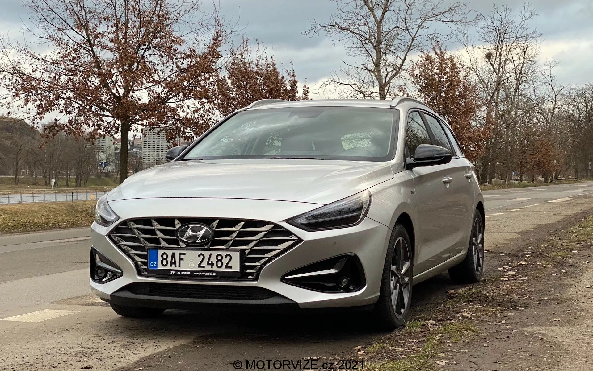 Obrázok predstavuje strieborný Hyundai i30 Kombi 2020 zachytený z nízkeho trojštvrťového uhla spredu, ukazujúci dizajn z pohľadu vodiča. Uhol zdôrazňuje veľkú kaskádovitú mriežku s výrazným logom Hyundai, moderné LED svetlomety, viacramenné zliatinové kolesá, integrované denné svetlá pod svetlometmi a viditeľný radarový senzor pre bezpečnostné funkcie. Zvýraznené sú aj aerodynamické línie a kontúry auta, so spätnými zrkadlami s integrovanými smerovkami. Pozadie zahŕňa obrubník, stromy a zamračené nebo, čo naznačuje prímestské vonkajšie prostredie.