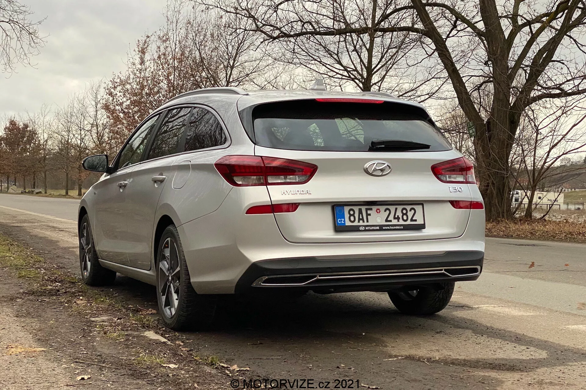 Zadný trojštvrťový pohľad na strieborný Hyundai i30 kombi z roku 2020, ktorý ukazuje dizajnové prvky zadnej časti vozidla, vrátane zadných svetiel s moderným dizajnom, ktoré môžu mať LED technológiu, zadného stierača, značiek auta a názvu modelu. Obrázok tiež zachytáva dva výfukové koncovky integrované do zadného nárazníka, zliatinové kolesá a čiastočný bočný profil, ktorý naznačuje dostatočný priestor v kabíne. Auto je zaparkované na dvojprúdovej ceste v vidieckej alebo polovidieckej oblasti s stromami a otvoreným priestorom v pozadí.