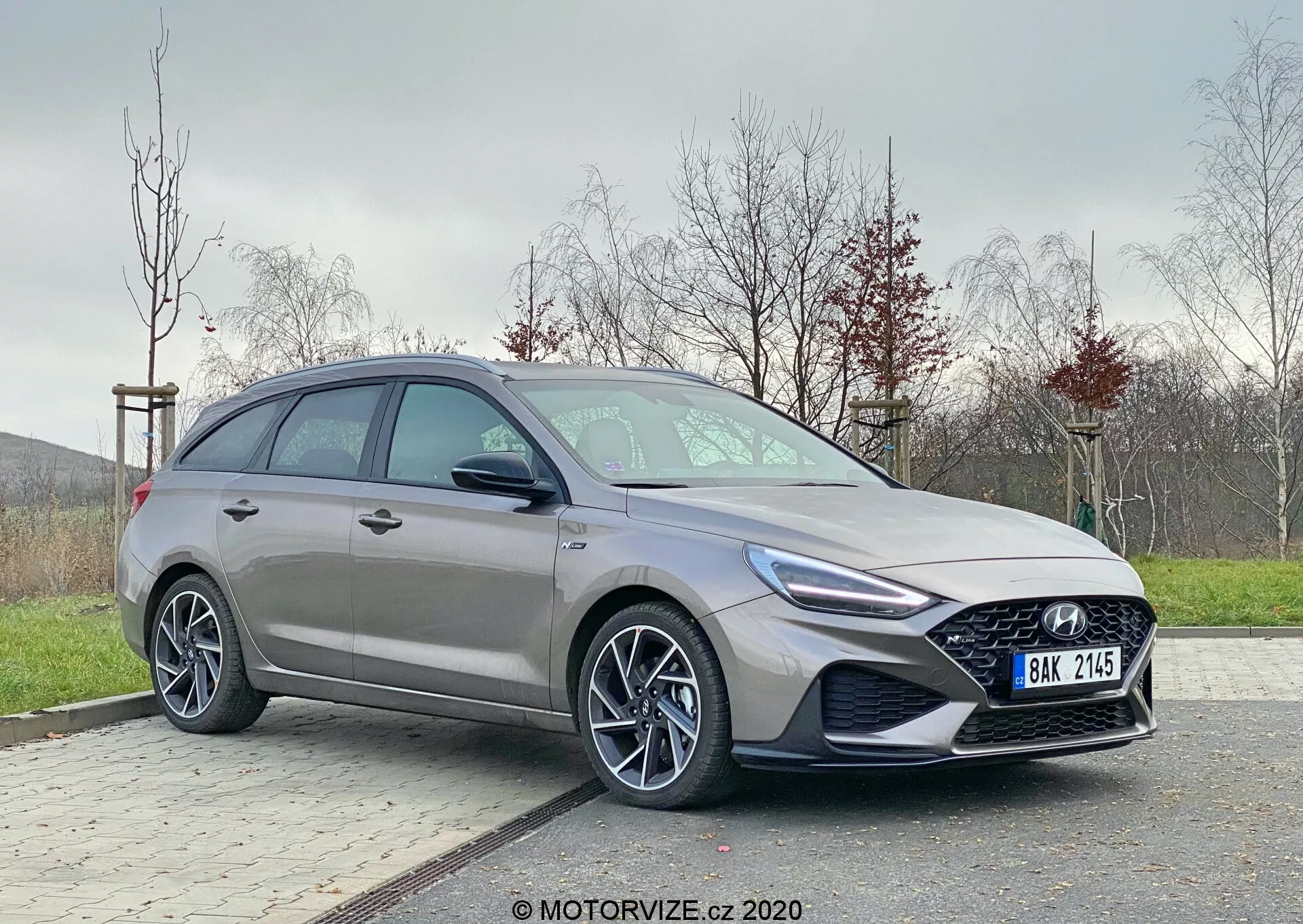 Moderný Hyundai i30 2020 z pohľadu trištvrť zpredu. Zameranie na vodičovu stranu ukazujúc elegantné dizajnové detaily ako agresívne svetlomety s LED dennými svetlami, výraznú mriežku chladiča, viacramenné zliatinové kolesá a kontúrový bočný profil. Umiestnený mimo centra na vydláždenej ploche s trávnatým pozadím, zobrazujúci dynamický, moderný hatchback s pokročilými automobilovými technológiami.