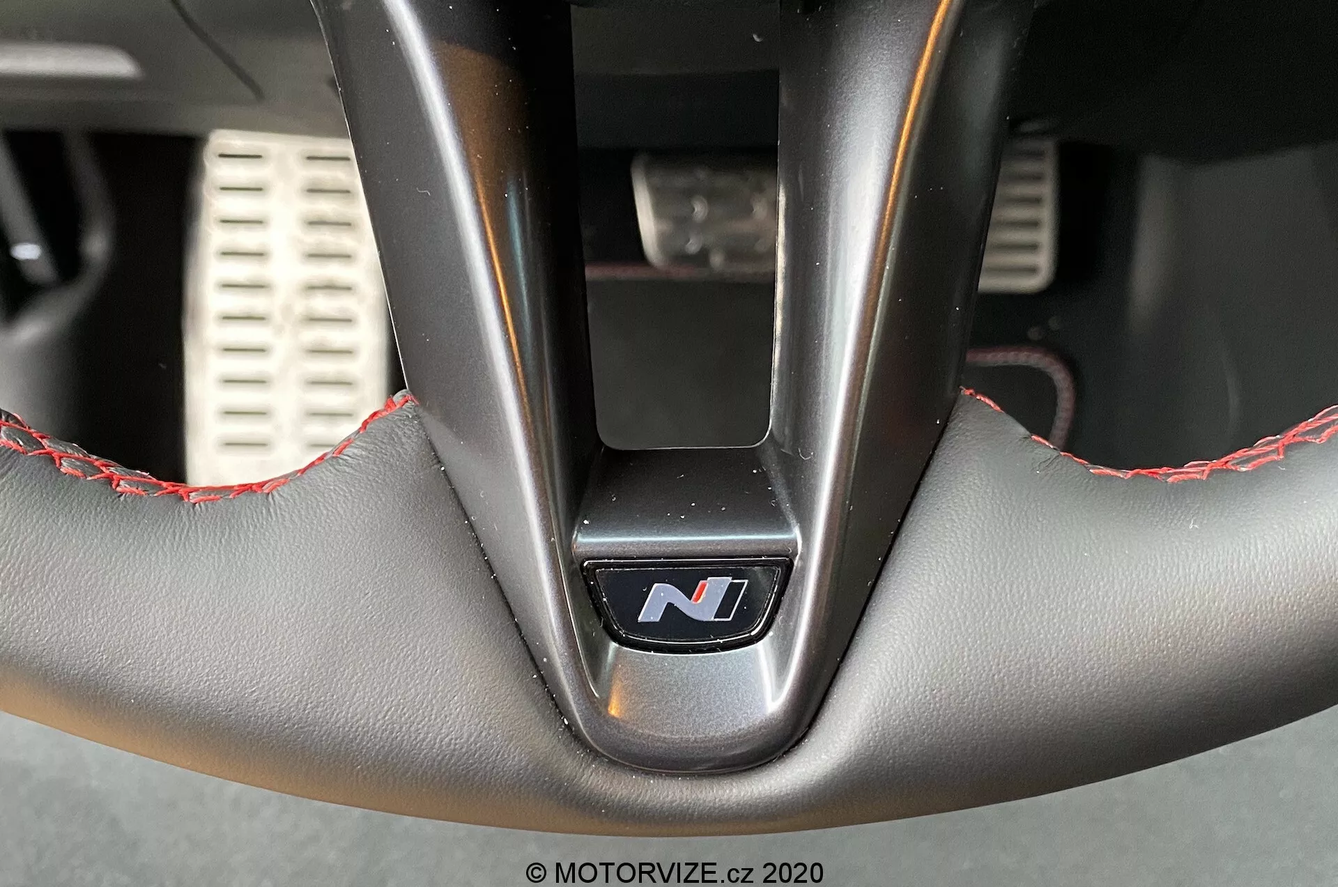 Detailný záber interiéru volantu Hyundai i30 2020 (i30-kombi) zospodu, zdôrazňujúci dolnú časť so stredovým logom 'N' poukazujúcim na výkonový variant, koženým čalúnením s červeným prešívaním, kovovými lúčmi a detailmi perforovaných športových pedálov v rozmazanom pozadí.