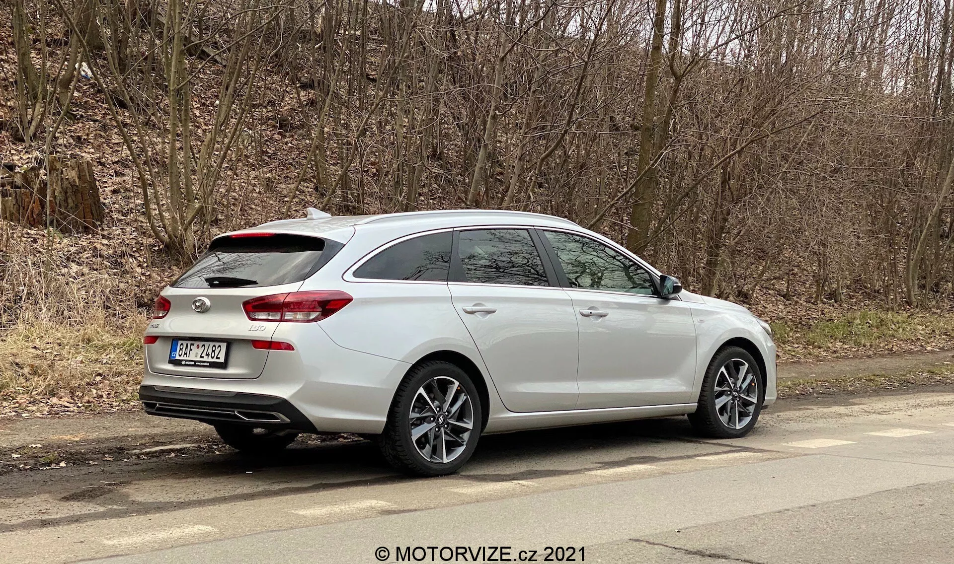 Hyundai i30 2020 kombi zaparkovaný na mestskej ceste s pohľadom na bočný profil a zadnú časť, so zadným strešným spojlerom, striebornými viacramennými zliatinovými kolesami, dvojitými výfukovými rúrami integrovanými do zadného nárazníka, zadnými svetlami zasahujúcimi do zadných dverí, strešnými lištami a bočnými zrkadlami s integrovanými smerovkami. Moderná technológia automobilu môže zahŕňať parkovacie senzory, zadnú kameru a LED zadné svetlá. Obrázok je dobre osvetlený a poskytuje detailný pohľad na dizajn a funkcie auta v mestskom prostredí.