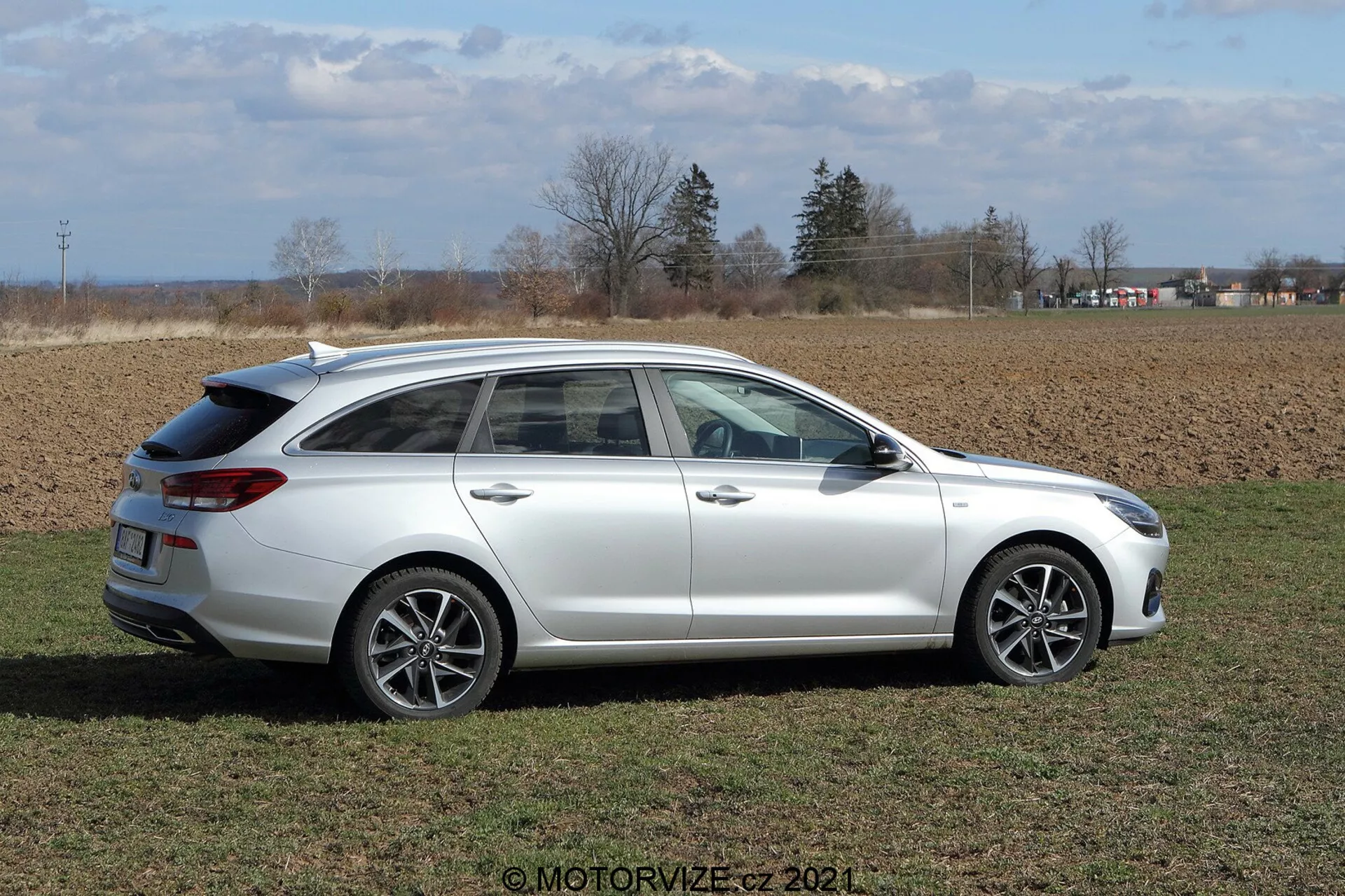 Bočný pohľad na Hyundai i30 Kombi z roku 2020 na dennom svetle na trávnatom poli. Auto má viacramenné zliatinové kolesá, strešné nosiče a zatmavené zadné okná, umiestnené vo vidieckom prostredí s poľnohospodárskymi poliami a čiastočne zamračenou oblohou v pozadí.