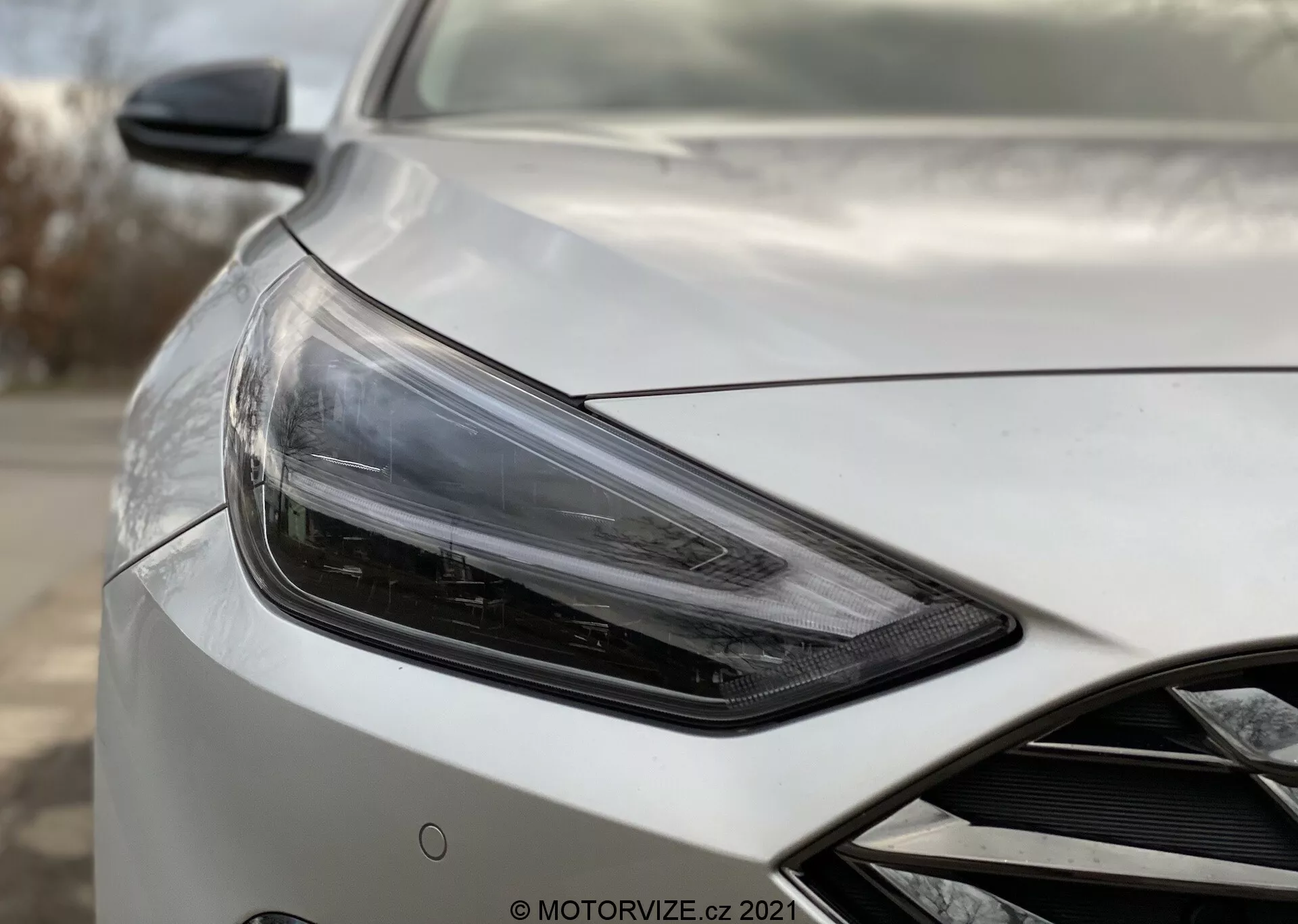Detailný záber na Hyundai i30 2020 Kombi, zobrazujúci prednú ľavú časť vozidla v uhle troch štvrtín smerom k vodičovi, so zameraním na elegantné, hranaté svetlomety s modernou LED alebo HID technológiou, doplnené o aerodynamické bočné zrkadlo s potenciálnymi pokročilými technologickými integráciami ako elektrické nastavenie a monitorovanie slepých uhlov. Karoséria vozidla má vysoko kvalitný perleťový alebo kovový povrch s aerodynamickými kontúrami na kapote a náznakom prednej mriežky. Pozadie je rozmazané, aby sa zdôraznili detaily vozidla.