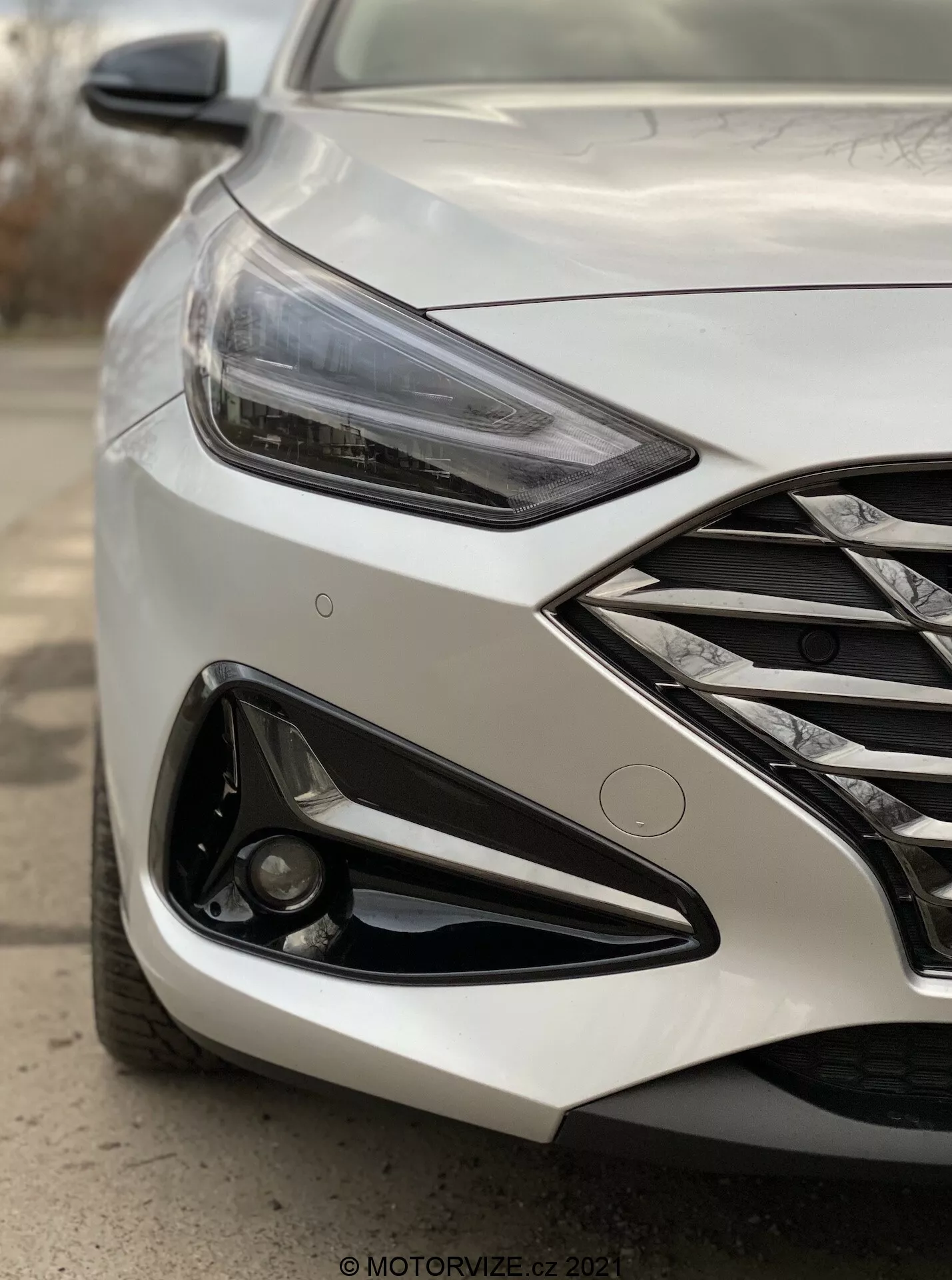 Zblízka uhlový záber prednej ľavej strany moderného Hyundai i30 Kombi 2020, zameriavajúci sa na svetlomet a oblasť predného nárazníka. Záber z nízkeho uhla pri prednom kolese zdôrazňuje ostrý uhlový LED svetlomet, mriežku s chrómovými prvkami, agresívny dizajn nárazníka s veľkým nasávacím otvorom a mlhovku zabudovanú v čiernom obložení. Metalický lak odráža svetlo a dizajnové línie na kapote zvýrazňujú dynamický vzhľad vozidla. Rozmazané pozadie zameriava pozornosť na detaily auta, naznačujúc pokročilé funkcie ako adaptívne osvetlenie a potenciálne parkovacie senzory.