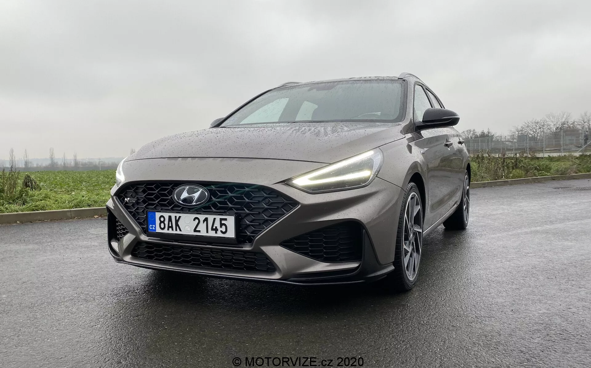 Obrázok zobrazuje moderný Hyundai i30 Kombi 2020 sedan s výrazným logom Hyundai na mriežke. Záber je zhotovený z nízkeho uhla, zdôrazňujúc trojštvrtečný pohľad na prednú časť vozidla a čiastočne na jeho bočný profil. Predná časť vozidla má veľkú čiernu mriežku, dynamické LED svetlomety a hmlovky alebo denné svetlá v dolných rohoch. Predný nárazník obsahuje stredný sací otvor a dekoratívne bočné otvory s športovým vzhľadom. Kapota má výrazné kontúrové línie. Vozidlo je v metalickej alebo grafitovej šedej farbe s mokrým povrchom, čo môže naznačovať dážď. Stierače sú v kľudovej polohe a mierne objemná základňa pri spätnom zrkadle môže naznačovať pokročilé senzory alebo kamery. Vozidlo je na mokrej ceste s neostro zobrazeným otvoreným pozadím.