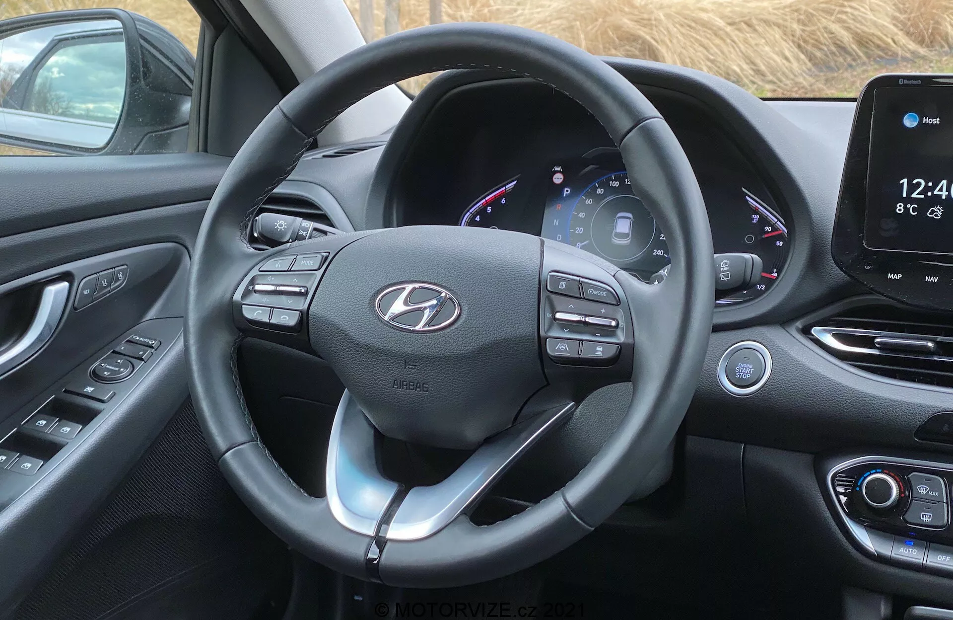 Obrázok interiéru prednej kabíny Hyundai i30 2020 z pohľadu vodiča. Zameriava sa na moderný volant s viacerými ovládačmi a pádlami radenia, s koženým obalom a viditeľným šitím a logom v strede. Za volantom je čiastočne digitálny prístrojový panel zobrazujúci rýchlomer, otáčkomer, merač paliva a iné informácie o vozidle s farebným rozhraním. Napravo je stredová konzola so systémom dotykovej infotainment obrazovky, ktorý zobrazuje čas, teplotu a domáce rozhranie, s fyzickými tlačidlami a kolieskami na ovládanie klimatizácie nižšie. Kabína sa môže pochváliť moderným dizajnom so zakrivenými povrchmi, mäkkými materiálmi a čierno-striebornou alebo chrómovou farebnou kombináciou, ktorá zvýrazňuje moderný a sofistikovaný vzhľad.