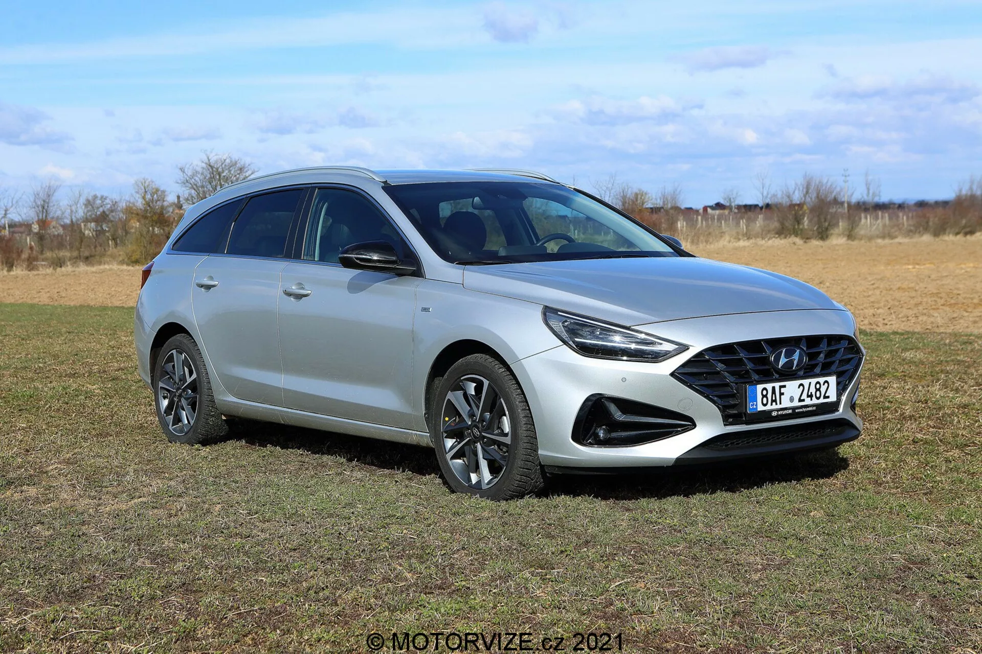 Hyundai i30 Kombi 2020 fotografovaný z ľavého predného trojštvrťového uhla na otvorenom poli. Elegantný strieborný hatchback s moderným aerodynamickým dizajnom, výraznou prednou mriežkou, ostrými LED svetlometmi, viaclúčovými zliatinovými kolesami, hmlovými svetlami, strešným spojlerom a strešnými lyžinami. Vonkajšie pozadie s pustými stromami zdôrazňuje pokročilú osvetľovaciu technológiu a možné bezpečnostné funkcie ako integrované senzory a smerovky na bočných zrkadlách.