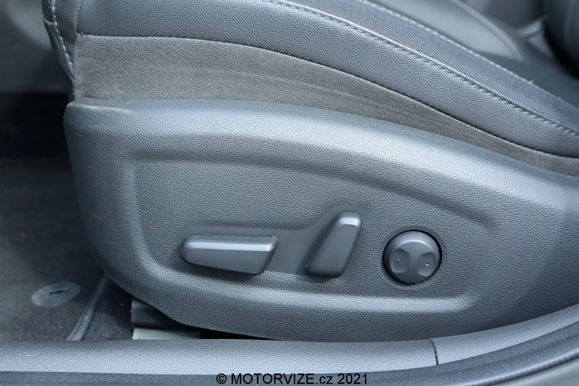 Detailný pohľad na interiér Hyundai i30 2020, zameraný na ovládanie sedadla vodiča z bočného uhla. Viditeľné funkcie zahŕňajú ovládanie nastavenia sedadla: predný bar-grip na posúvanie, stredná páka na sklopenie a zadné okrúhle ovládanie na nastavenie výšky. Plastový kryt sa integruje so štýlom bočnej časti sedadla a čalúnenie z tkaniny poskytuje odolnosť a pohodlie.
