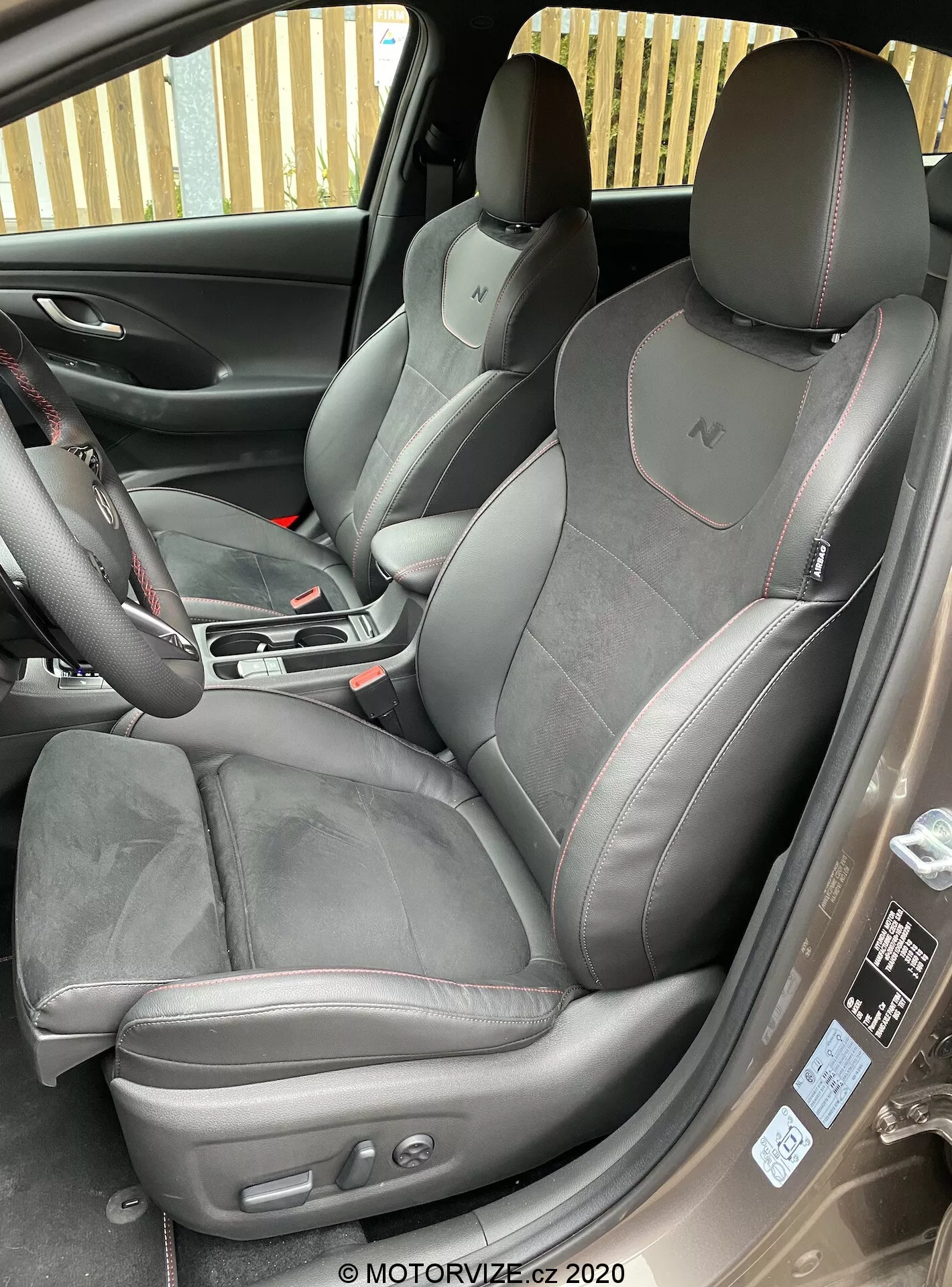 Tento detailný obrázok zobrazuje interiér prednej kabíny Hyundai i30 2020 (i30-kombi) z mierne zvýšeného uhla mimo otvorených predných dverí spolujazdca. Otvorené dvere odhaľujú bočnú stranu prémiových športových sedadiel, ktoré sú čalúnené semišom alebo alcantarou a kožou s kontrastným prešívaním. Integrované opierky hlavy s logom 'N' naznačujú úroveň výbavy vozidla. Sedadlo vodiča obsahuje tlačidlá nastaviteľné elektricky na nastavenie výšky a pomoci elektrických ovládí 
