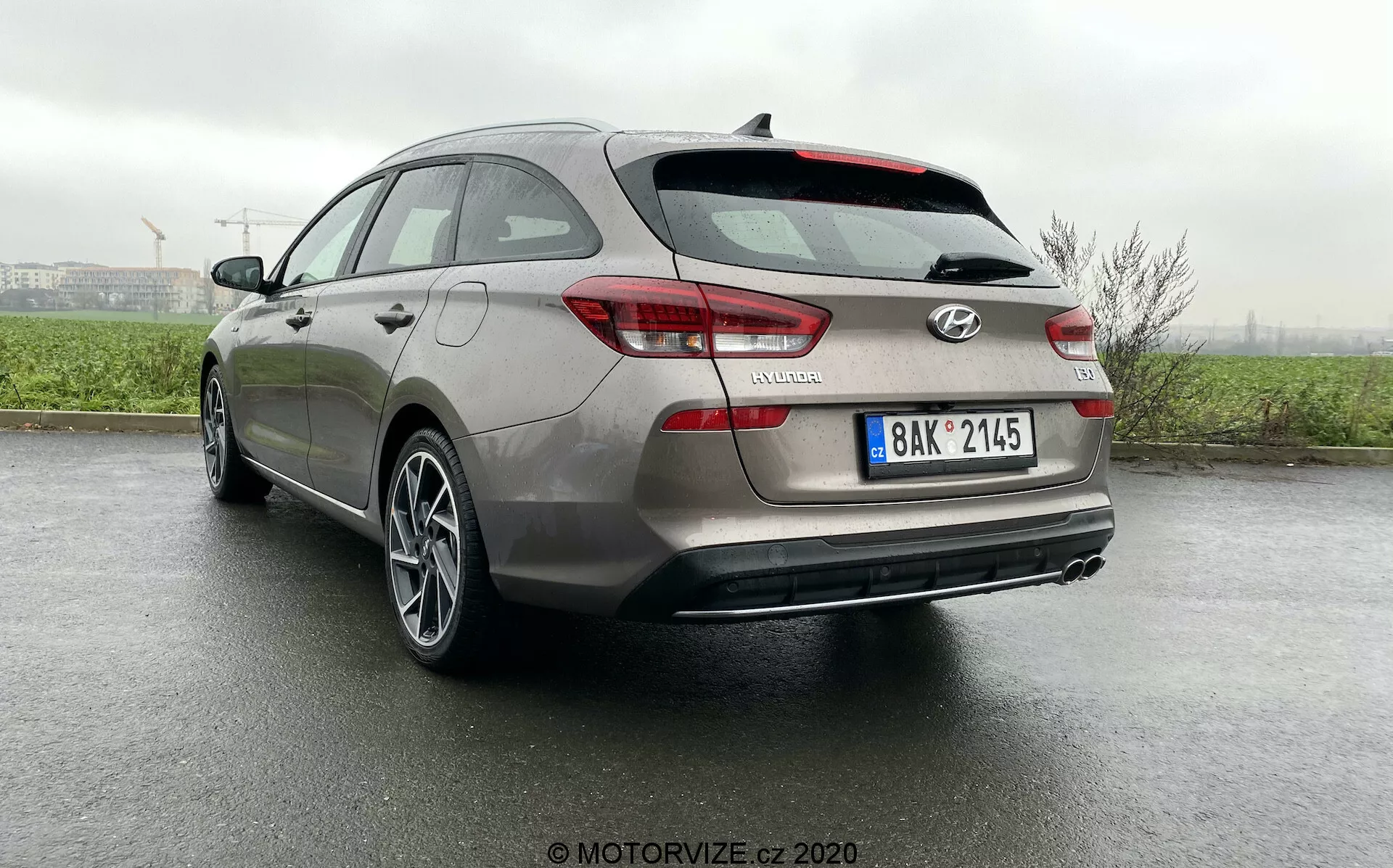 Zadný trojštvrtečný pohľad na Hyundai i30 Kombi 2020 z nízkeho uhla, ktorý zobrazuje detaily dizajnu vrátane zadných svetiel, zadného nárazníka, dverí kufra, zadnej parkovacej kamery, zliatinových kolies a odraziek na mokrom povrchu po nedávnom daždi, zdôrazňujúc pokročilé systémy asistencie vodiča a estetický vzhľad vozidla.