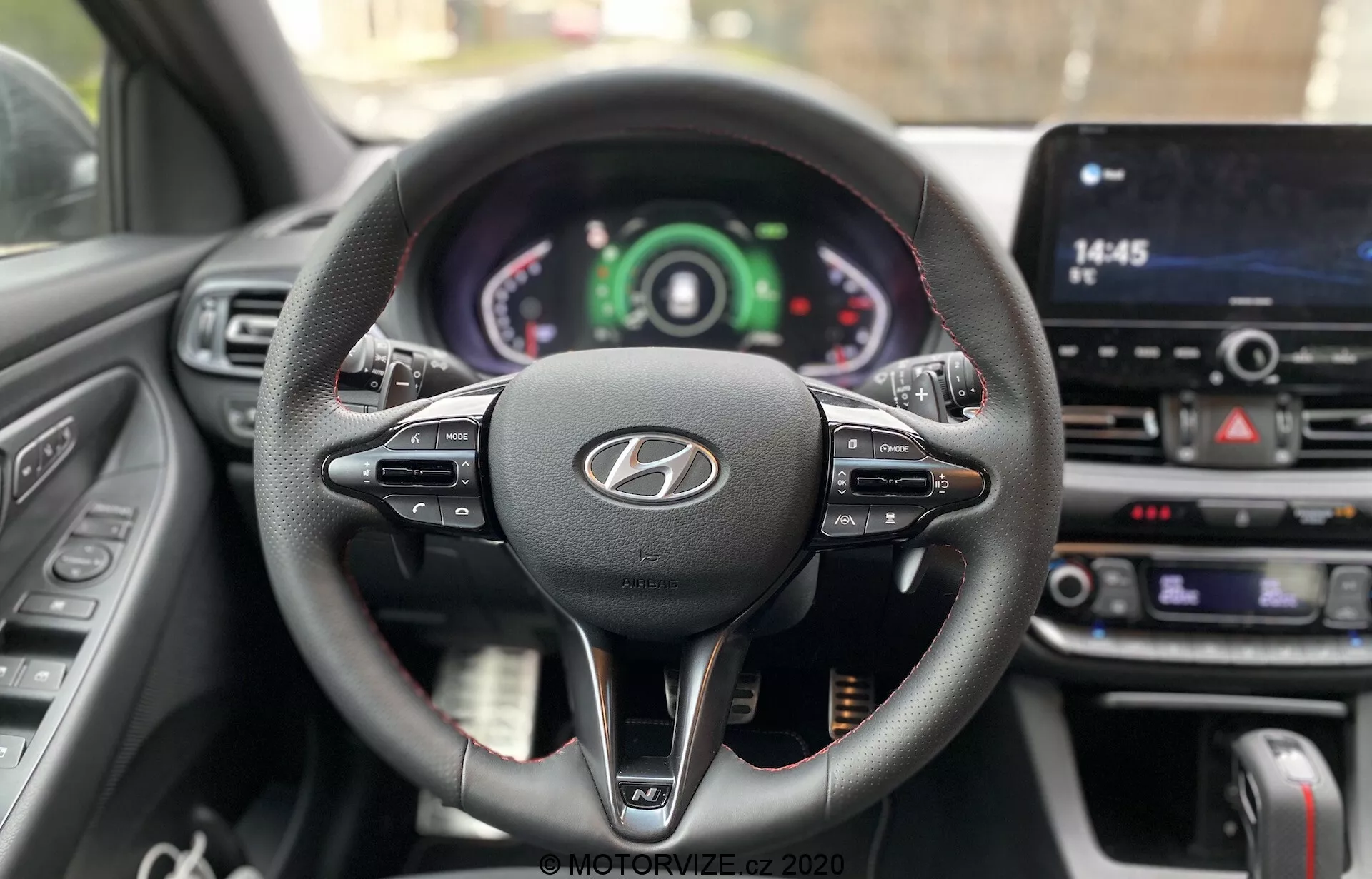 Pohľad z perspektívy vodiča na volant a prístrojovú dosku Hyundai i30 2020 s trojramenným koženým dizajnom s multifunkčnými ovládačmi, výrazným logom Hyundai a prístrojovým panelom s analógovými a digitálnymi displejmi. Centrálne umiestnený dotykový displej infotainmentu a tlačidlá klimatizácie zvýrazňujú pokročilé technológie v aute a interaktívne prvky vodiča.