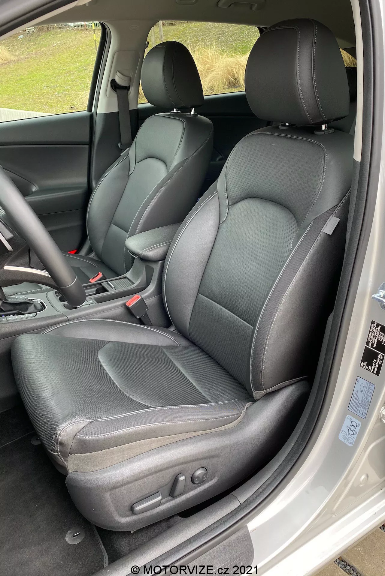Detailný pohľad na prednú časť interiéru Hyundai i30 Kombi 2020, zameraný na stranu vodiča. Perspektíva je mierne nad úrovňou sedadla, diagonálne nadol do kabíny, ako keby ste vstupovali z zadných dverí. Ľavé kožené sedadlo vodiča je výrazne zobrazené s viditeľným prešívaním a elektronickými ovládačmi nastavenia na bočnej strane. Bezpečnostný pás a ľavá tretina trojramenného volantu sú viditeľné. Čiastočne sú viditeľné časti stredovej konzoly, vrátane zvislej automatickej páky radenia v polohe ‚parkovanie‘, a časti prístrojovej dosky a výduchu vzduchu. Kabína má čierny monotónny farebný schéma, s rôznymi textúrami na sedadlách, dverových paneloch a prístrojovej doske. Fotografia je dobre osvetlená prirodzeným denným svetlom, čo naznačuje zatienené alebo zamračené prostredie.