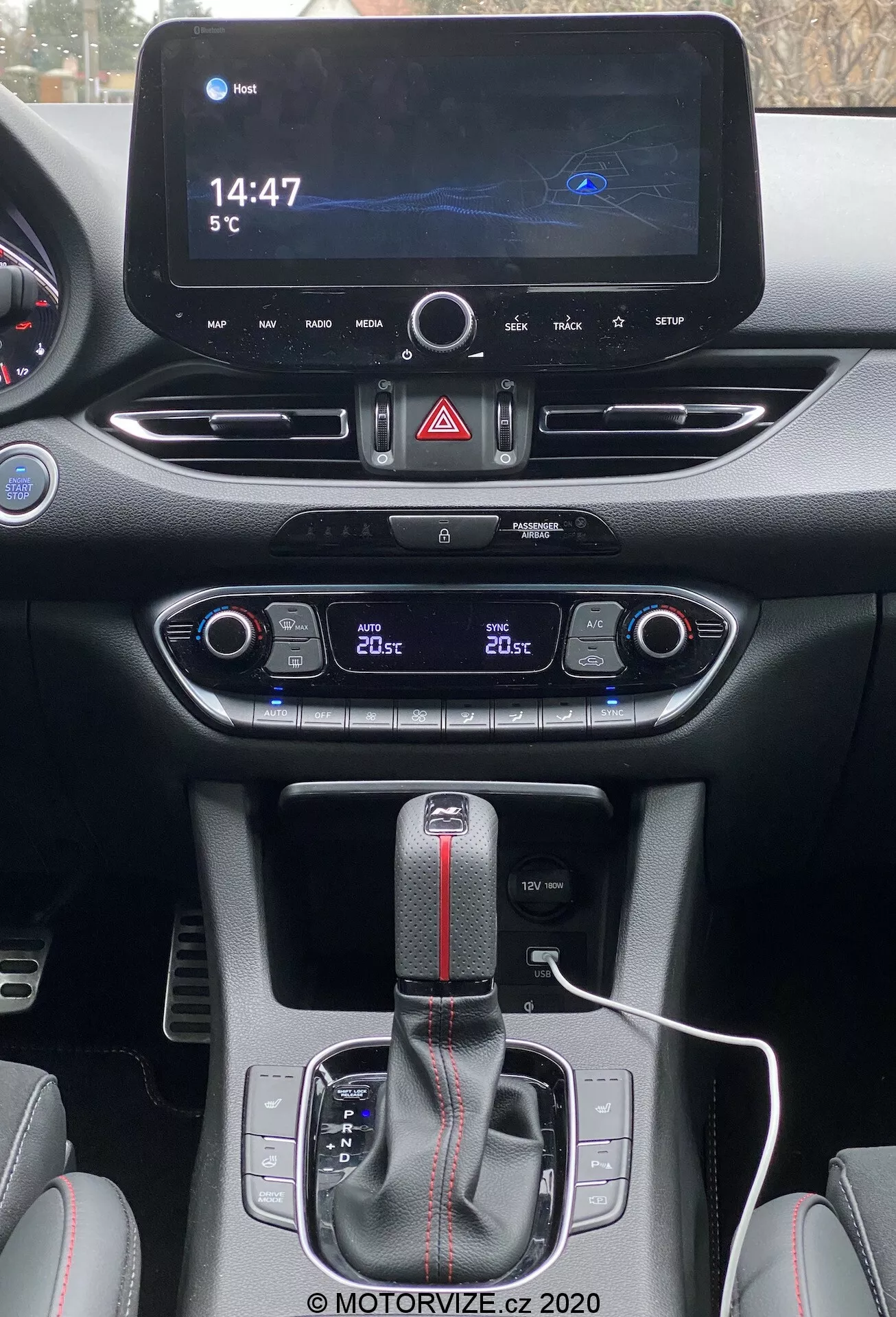Pohľad vodiča na centrálnu konzolu Hyundai i30 Kombi 2020. Nahoře moderný infotainment displej zobrazuje mapu navigácie, digitálne hodiny s časom '14:47' a okolnú teplotu '5°C'. Dole sú tlačidlo výstražných svetiel a vetracie otvory. Nižšie ovládací panel klimatizácie s dvojzónovým systémom ukazuje '20°C' pre obe strany. Automatická radiaca páka s čiernym krytom a červeným pruhom naznačuje športový úpravu. Biely nabíjací kábel je zapojený v blízkosti radiacej páky, obklopený ovládacími prvkami pravdepodobne pre jazdné režimy alebo trakciu. Materiály ukazujú kombináciu hladkých a štruktúrovaných povrchov s kontrastným prešívaním, zdôrazňujúc moderný, športový interiér.
