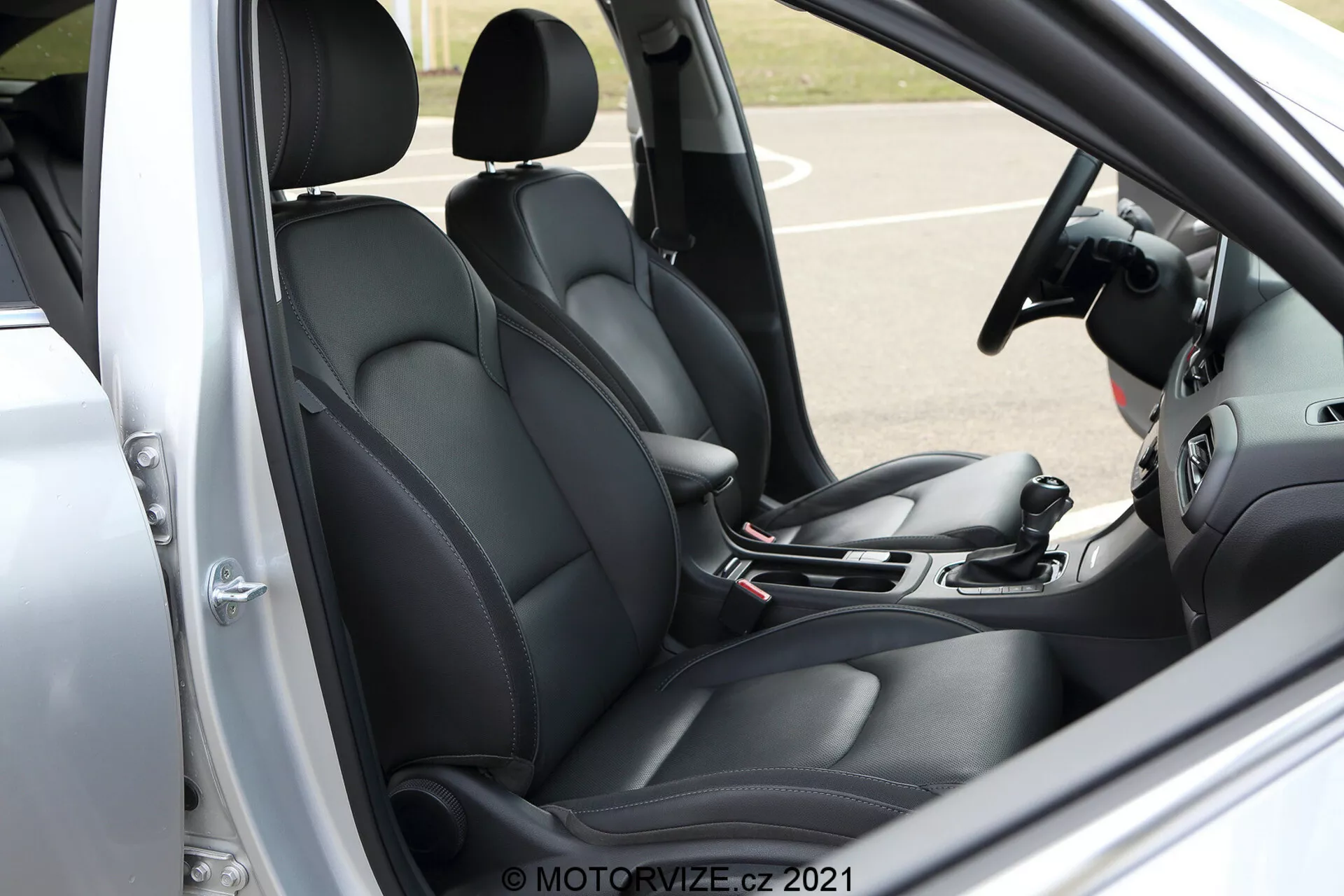 Detailný pohľad z boku na prednú kabínu Hyundai i30 Kombi 2020. Obrázok zobrazuje vodičovo sedadlo a časť spolujazdcovho sedadla, obidve čalúnené v čiernej farbe s kontúrovaným dizajnom pre väčšie pohodlie a oporu. Viditeľné z vodičovej strany s otvorenými dverami, scéna zahŕňa radiacu páku, ručnú brzdu a časť infotainment systému a systému klimatizácie, čo naznačuje moderné technologické prvky. Panel vodičových dverí, zobrazujúci ovládače elektrických okien a možno nastavenie zrkadiel, pridáva k celkovému funkčnému dizajnu. Volant, aj keď čiastočne orezaný, naznačuje integrované ovládacie prvky pre lepšiu interakciu s vozidlom. Vodotlač 'MOTORVIZE.CZ 2021' v pravom dolnom rohu.