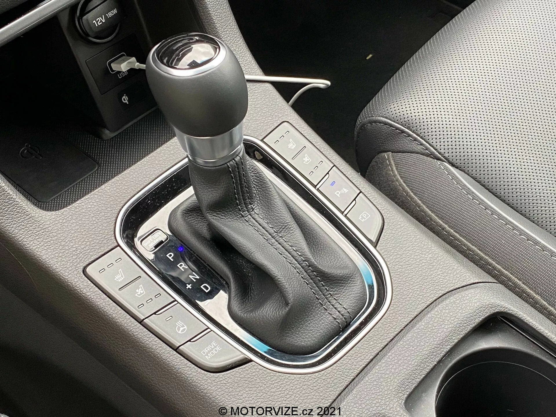 Detailný záber na automatickú prevodovku Hyundai i30 2020, foto pod uhlom 45 stupňov. Radiaca páka je obklopená lesklou čiernou úpravou s koženým krytom a viditeľným šitím. PRND schéma je jasne viditeľné spolu s indikáciami manuálneho alebo športového režimu. Tlačidlá naľavo ovládajú jazdné režimy, zatiaľ čo na pravej strane sú ovládacie prvky stability alebo trakcie vozidla. Je viditeľná aj elektronicky ovládaná parkovacia brzda. Dobre osvetlený obrázok zdôrazňuje vysoko kvalitné a precízne spracovanie stredovej konzoly.