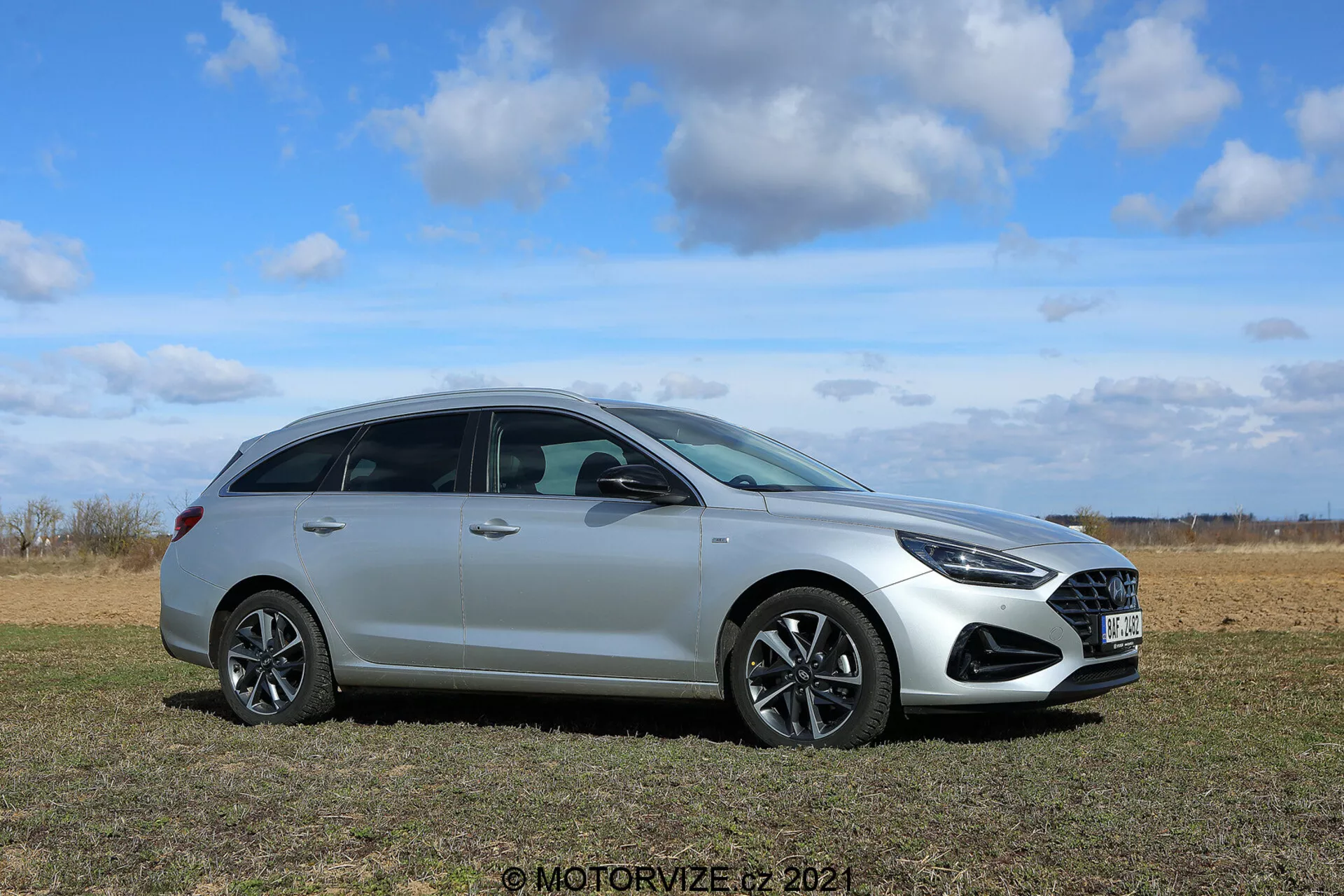 Hyundai i30 2020 Kombi - Moderný hatchback v trávnatej oblasti s jasnou oblohou v pozadí, bočný profil zvýrazňujúci prednú masku, dizajn kolies, svetlomety, bočné zrkadlá, okná, kľučky dverí a líniu strechy; vyniká elegantným metalickým lakom, zliatinovými kolesami a pokročilými svetelnými a aerodynamickými funkciami.