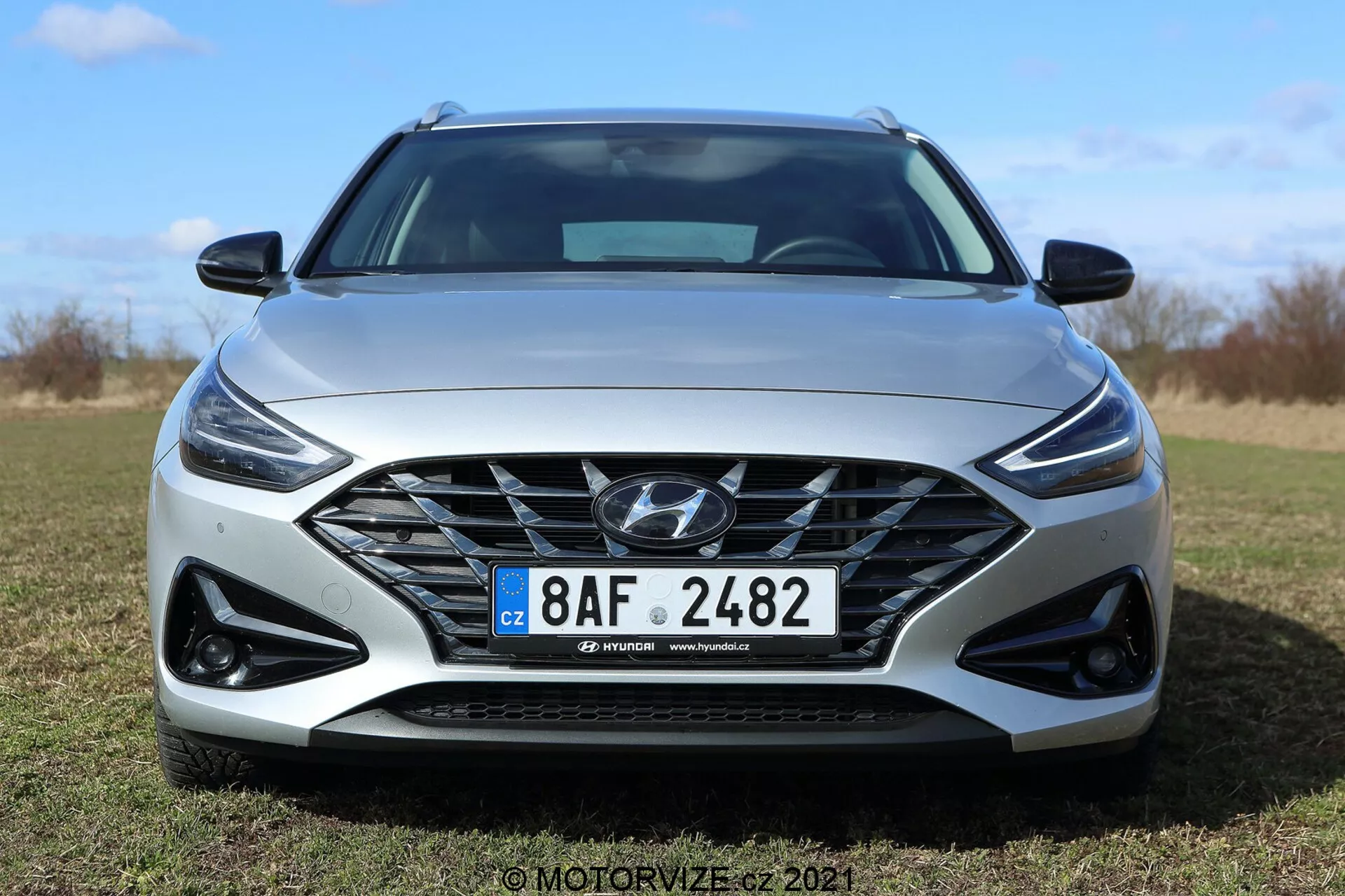 Predný pohľad na Hyundai i30 Kombi 2020 za jasného dňa v exteriérovom prostredí. Moderný dizajn auta obsahuje veľkú masku chladiča so zložitou mriežkou a logom Hyundai, elegantné LED svetlomety s dennými svetlami a športové kryty hmloviek v čiernej farbe. Dynamický predný nárazník a tvarovaná kapota zdôrazňujú súčasnú a aerodynamickú estetiku. Viditeľná je európska poznávacia značka a pravdepodobne aj zliatinové kolesá. Svetlošedá farba auta zvýrazňuje jeho sofistikovaný štýl.