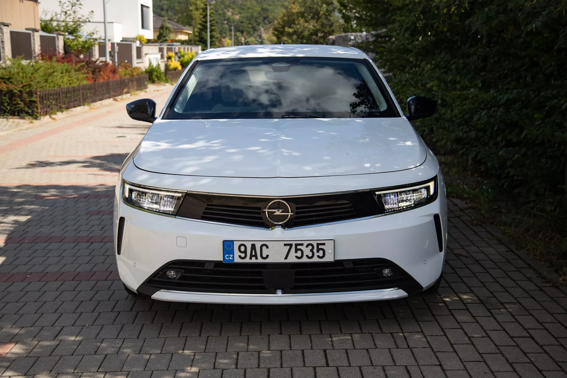 Predný pohľad na Opel Astra 2022 ukazuje moderný dizajn elektrického vozidla s LED svetlometmi, dekoratívnou mriežkou a aerodynamickým tvarom, zaparkované na príjazdovej ceste k rodinnému domu