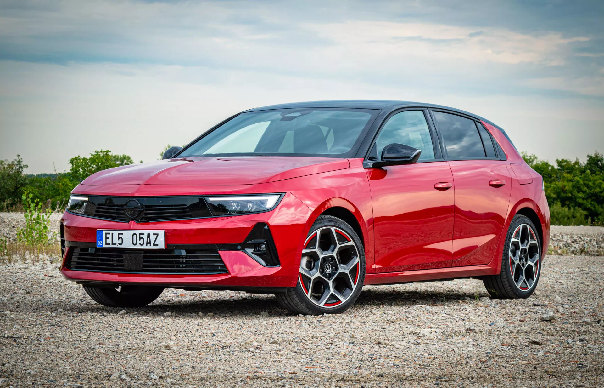 Červený hatchback Opel Astra 2022 fotografovaný z trojštvrťového predného pohľadu ukazujúci LED svetlomety, výraznú prednú mriežku, disky z ľahkých zliatin s viacerými ramenami a elegantný bočný profil na štrkovej povrchu s rozptýleným slnečným svetlom osvetľujúcim moderné funkcie auta