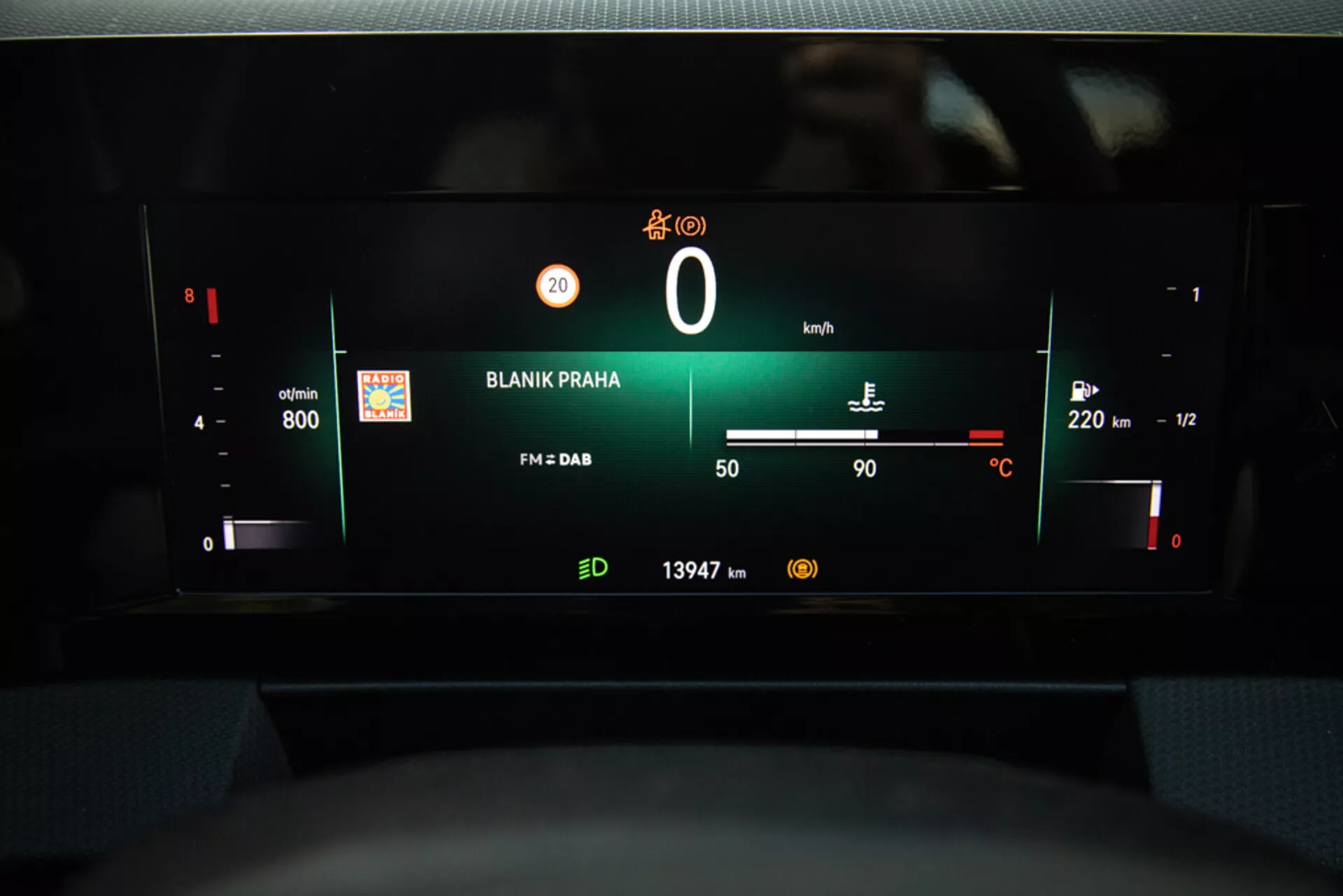 Pohľad na digitálnu palubnú dosku Opel Astra 2022 so zameraním na prístrojový panel s otáčkomerom, úrovňou batérie, rýchlosťou, ukazovateľom paliva, vonkajšou teplotou, rádiovou stanicou 'BLANIK PRAHA' a čítaním tachometra z pohľadu vodiča.