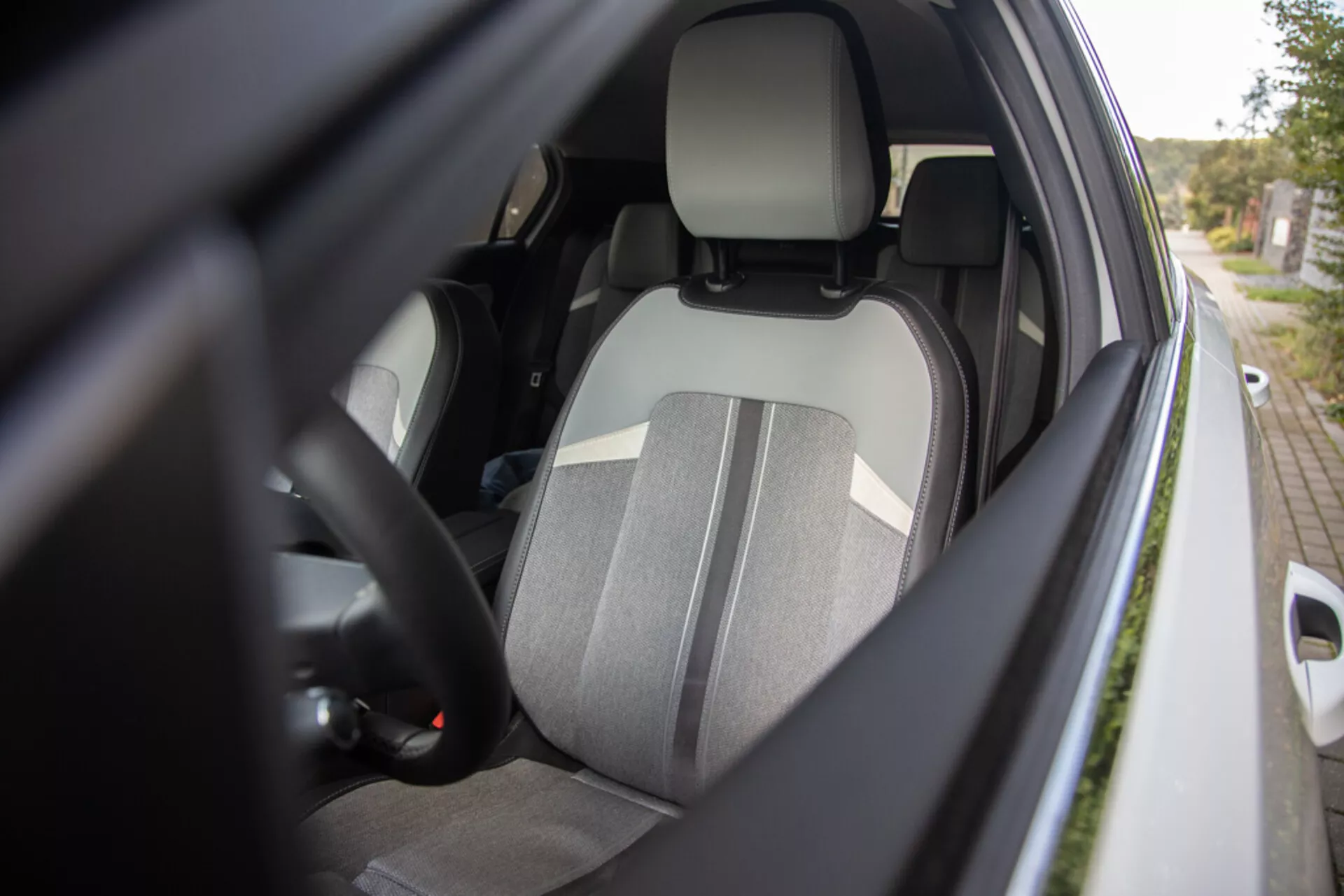 Detailný pohľad na interiér vodičskej strany Opel Astra 2022 zo stranného uhla zvonka ukazuje sedadlo vodiča s dvoma tónmi látky, integrovanú opierku hlavy, časť volantu s ovládacími prvkami, náhľad na prístrojovú dosku, zapnutý bezpečnostný pás, vnútro dverí s potenciálnymi ovládačmi elektrických okien a kryt bočného zrkadla v farbe karosérie s dôrazom na ergonómiu, bezpečnosť a pohodlie.