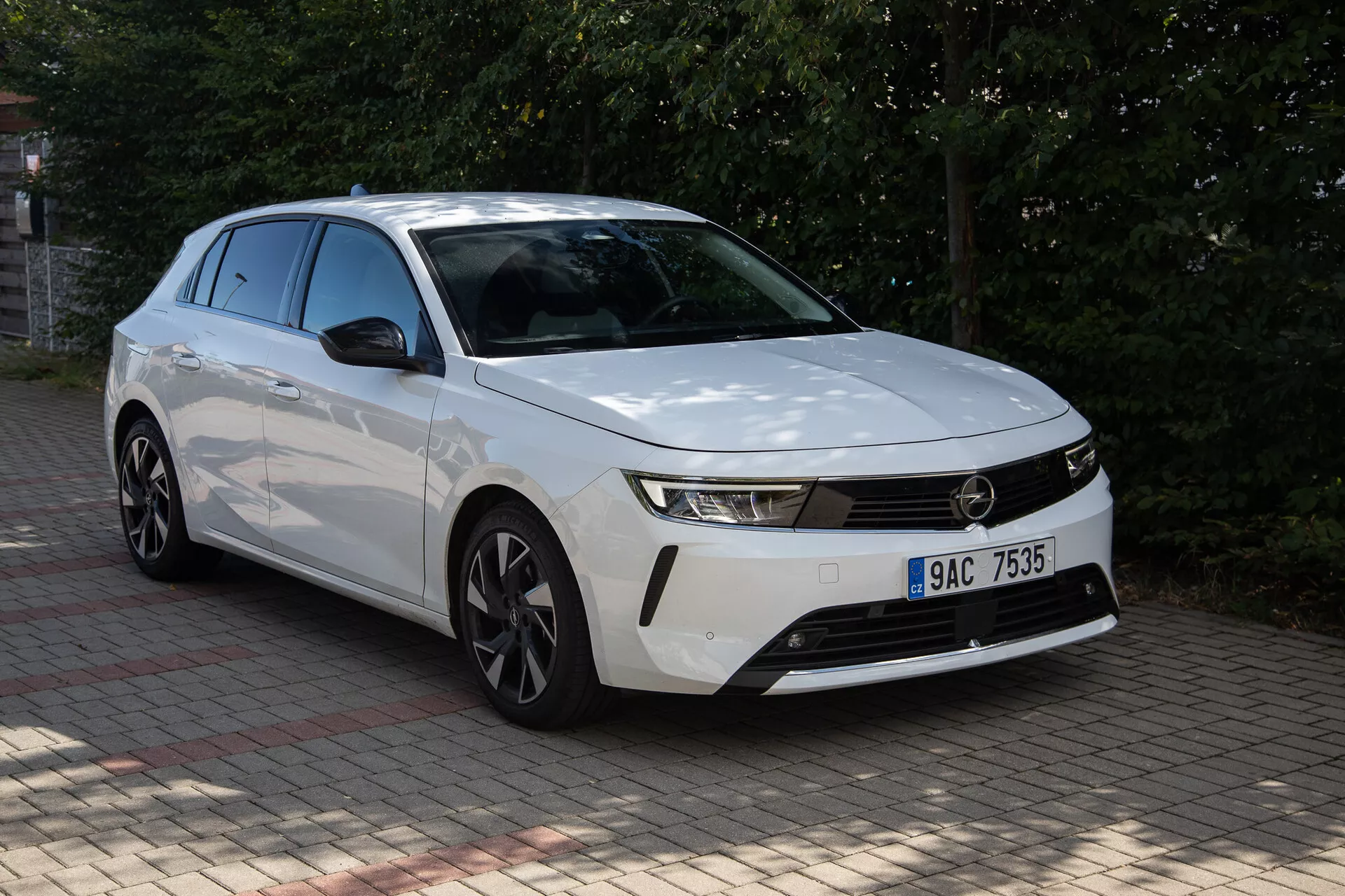 Biela Opel Astra 2022 s výrazným dizajnom prednej časti, štíhlymi svetlometmi, horizontálnou mriežkou, aerodynamickým nárazníkom, tvarovanou kapotou a disky z ľahkej zliatiny zachytená z trojštvrťového predného pohľadu vonku počas dňa