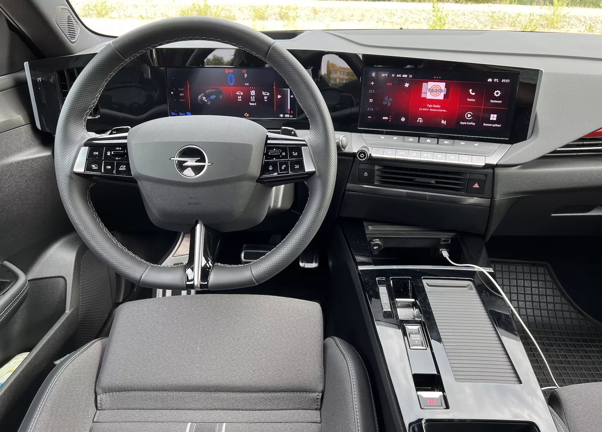 Interiér Opel Astra 2022 z pohľadu zo zadného sedadla, zobrazujúci trojramenný volant s integrovanými ovládačmi, digitálny prístrojový panel, dotykový displej infotainmentu, stredovú konzolu so zařazením a úložným priestorom, a kombináciu materiálov z látky a kovového efektu