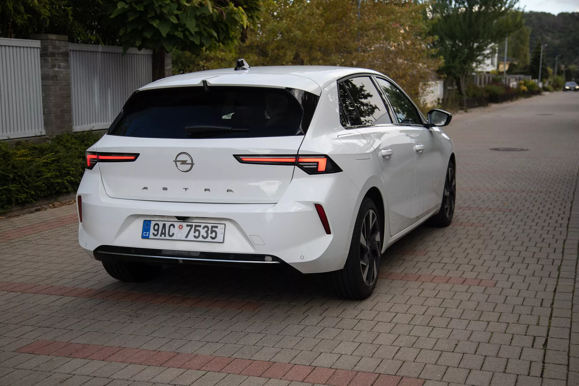 Moderný sedan Opel Astra 2022 v bielej farbe, zaparkovaný na povrchu z prepojených tehál, zobrazuje zozadu trojštvrťový uhol s dôrazom na vysoko technologické LED zadné svetlá, zadnú kameru, parkovacie senzory, čiastočný bočný profil vrátane pravej zadnej kolesa a dverí, spojený horizontálnym svetelným pásom cez veko batožinového priestoru, vybavený žraločou plutvou anténou pre zlepšenú konektivitu, vhodný na ilustráciu dizajnu zadnej časti vozidla a moderne automobilovej technológie