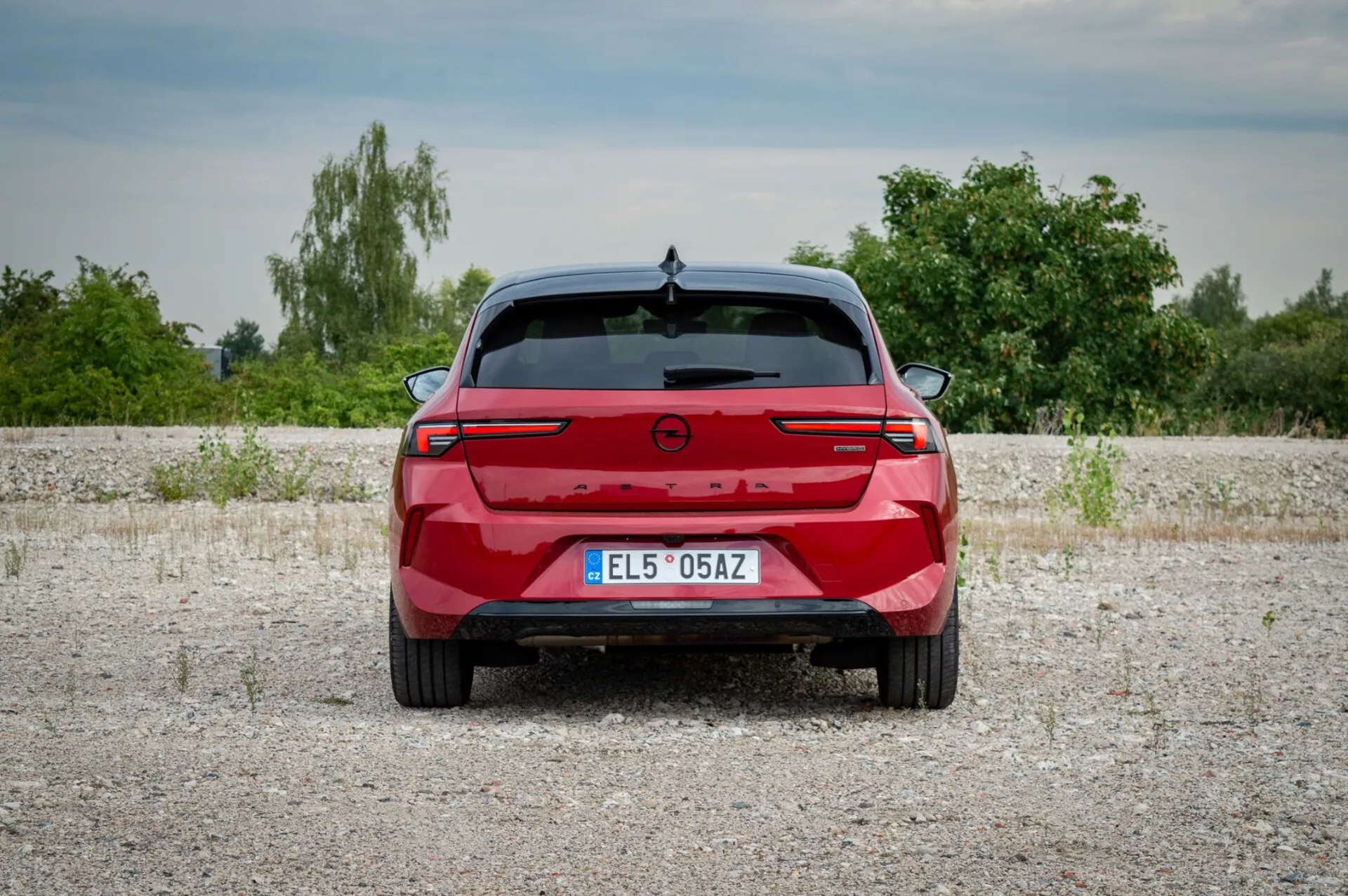 Opel Astra 2022 fotografovaná zozadu na štrkovej ploche ukazujúca zadné svetlá s pripájacou svetelnou lištou, parkovacie senzory a absenciu výfukových portov naznačujúcu potenciálny elektrický alebo hybridný model, na pozadí vegetácie a mierne zamračeného neba.