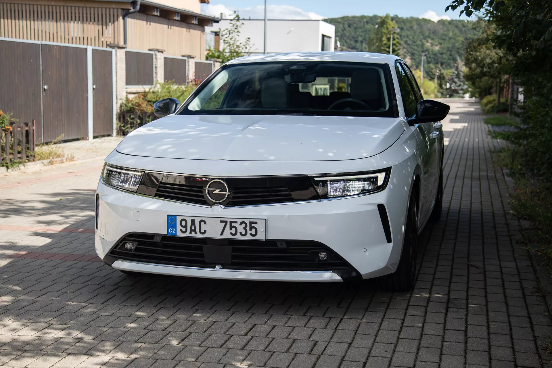 Pohľad z prednej trojštvrte na Opel Astra 2022 s LED svetlometmi, minimalistickým dizajnom mriežky, čiastočne viditeľnými zliatinovými kolesami a hladkými karosárskymi panelmi na európskej obytnej ulici