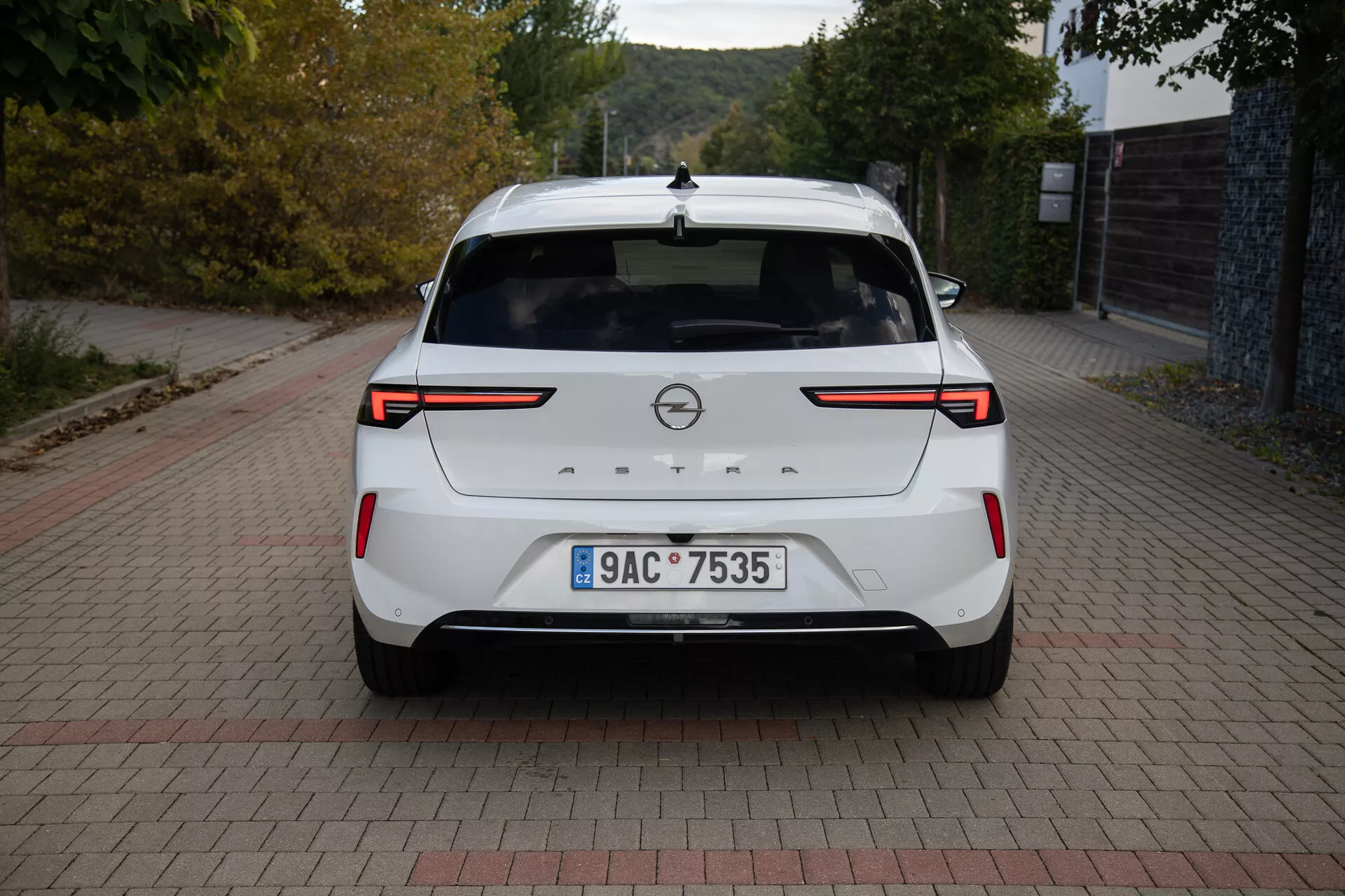 Zadný pohľad na biely Opel Astra 2022 ukazujúci LED zadné svetlá, parkovacie senzory/kameru v zadnej časti, značkový odznak, evidenčnú značku, zadný spojler, vysoko umiestnenú stop svetlo, anténa v tvare žraločej plutvy a čistý povrch laku, zaparkovaný na povrchu s tehlovým vzorom pri dennom svetle.